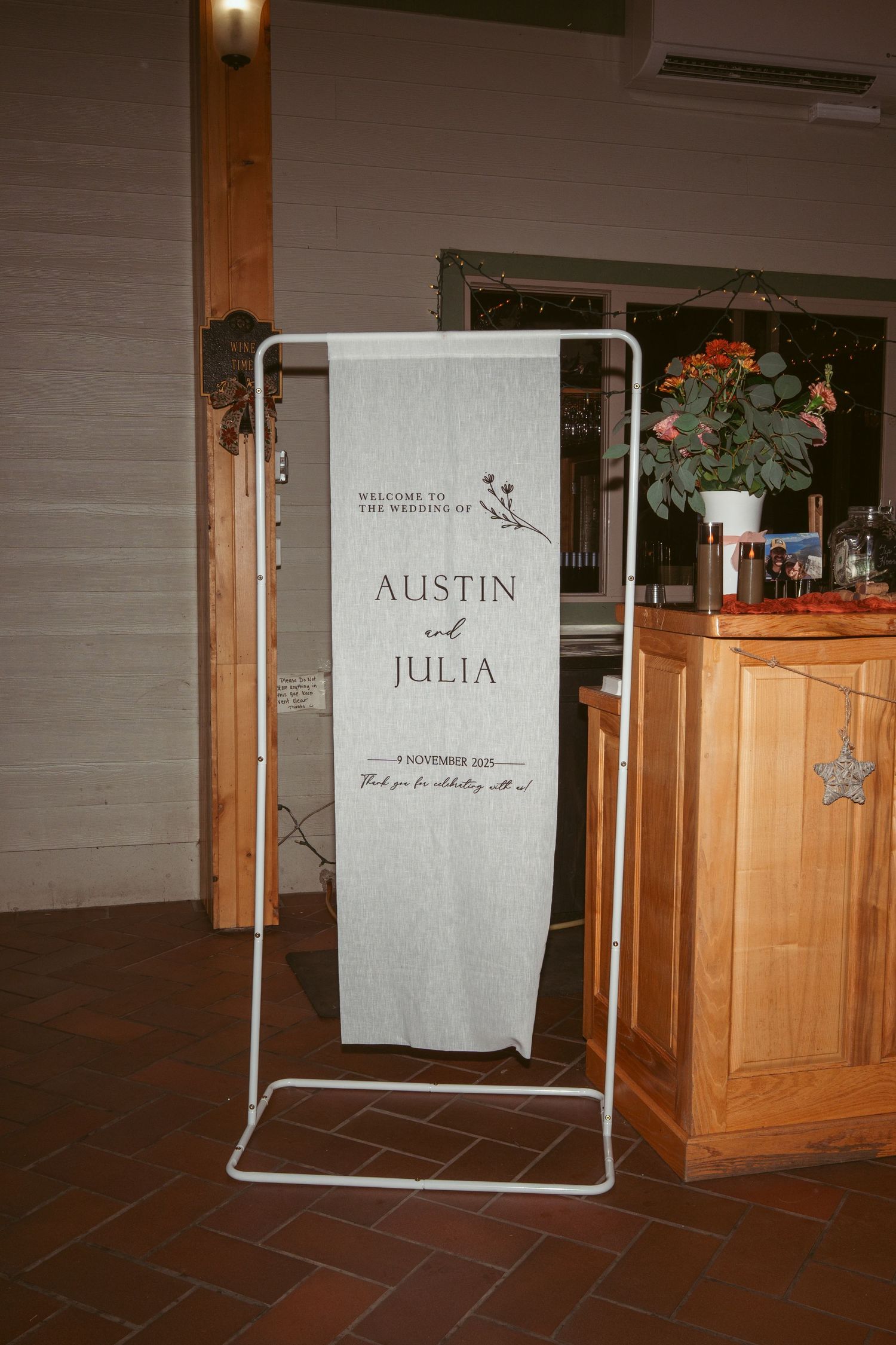 Wedding welcome sign on white fabric displayed on metal stand next to wooden cabinet.