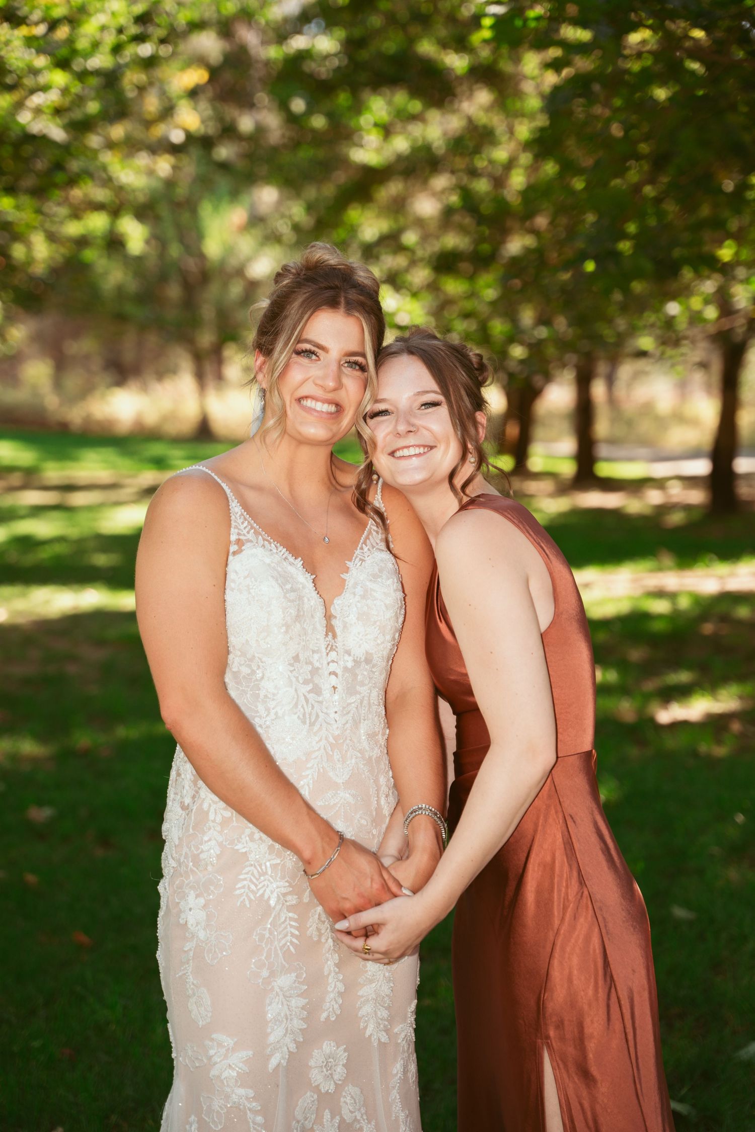 Two women embrace and smile outdoors, one wearing a lace wedding dress and the other in a rust-colored formal dress.