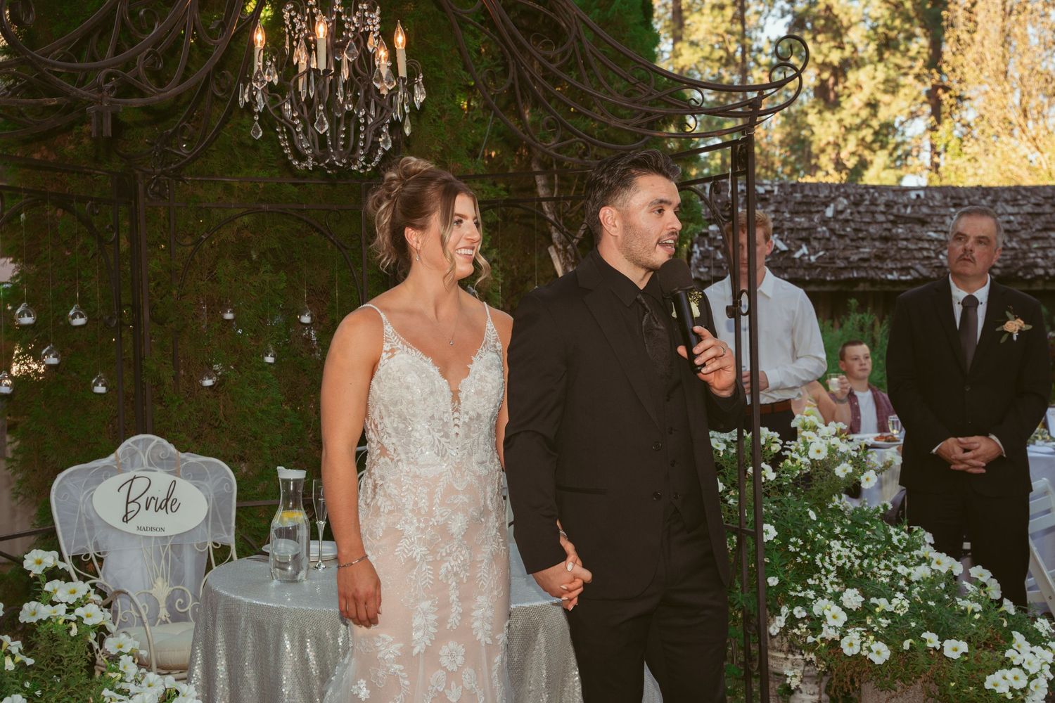 Elegant outdoor evening reception with hanging chandeliers and string lights among trees.