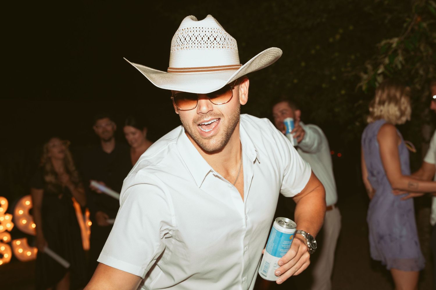 Party guests in casual attire dancing and socializing at a dimly lit outdoor event with cowboy hats.