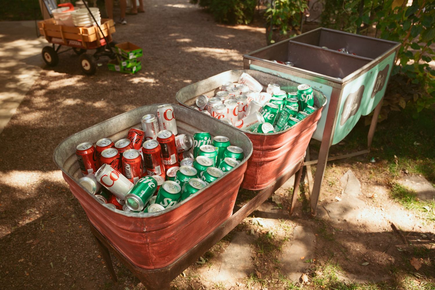 Red coolers filled with assorted soda cans arranged outdoors in dappled sunlight.