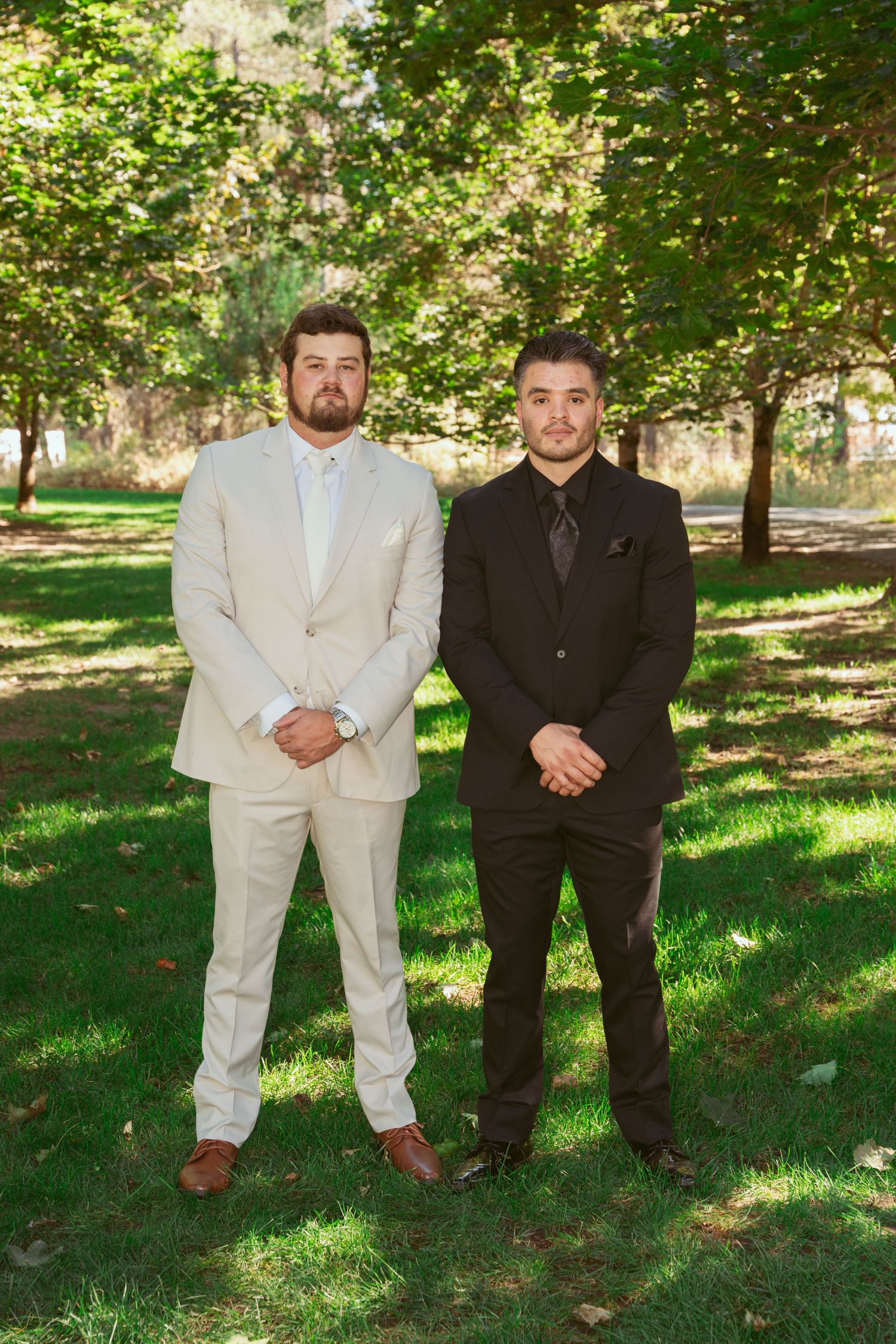 Groomsmen in contrasting black and white suits pose together on grass under shaded trees.