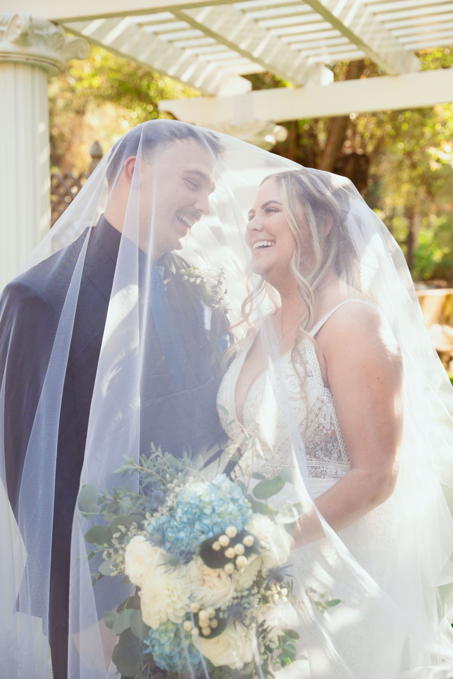 Joyful wedding moment captured under white pergola with couple sharing laughter beneath flowing veil.