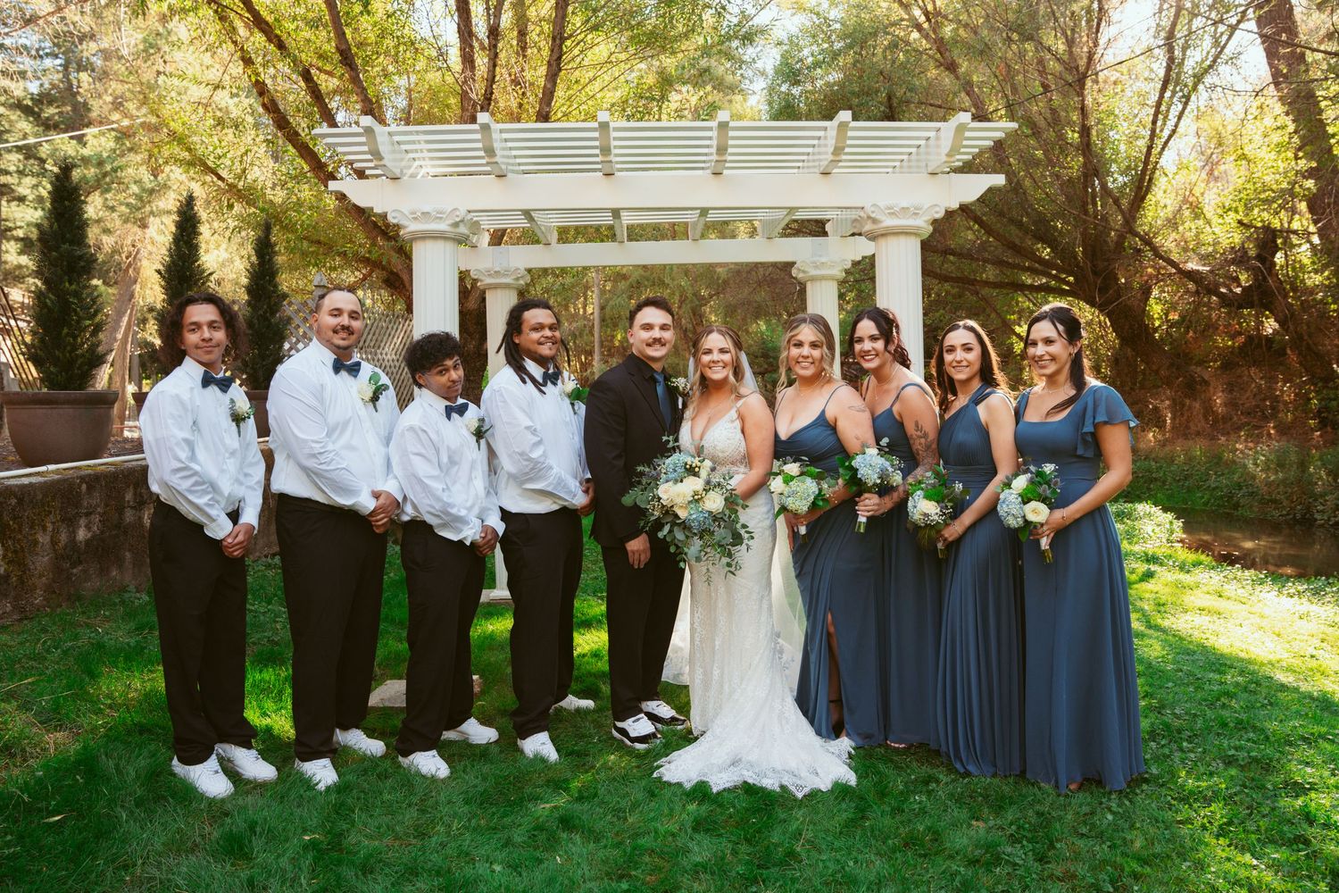 Wedding party in navy and white attire posed under white pergola in garden setting.