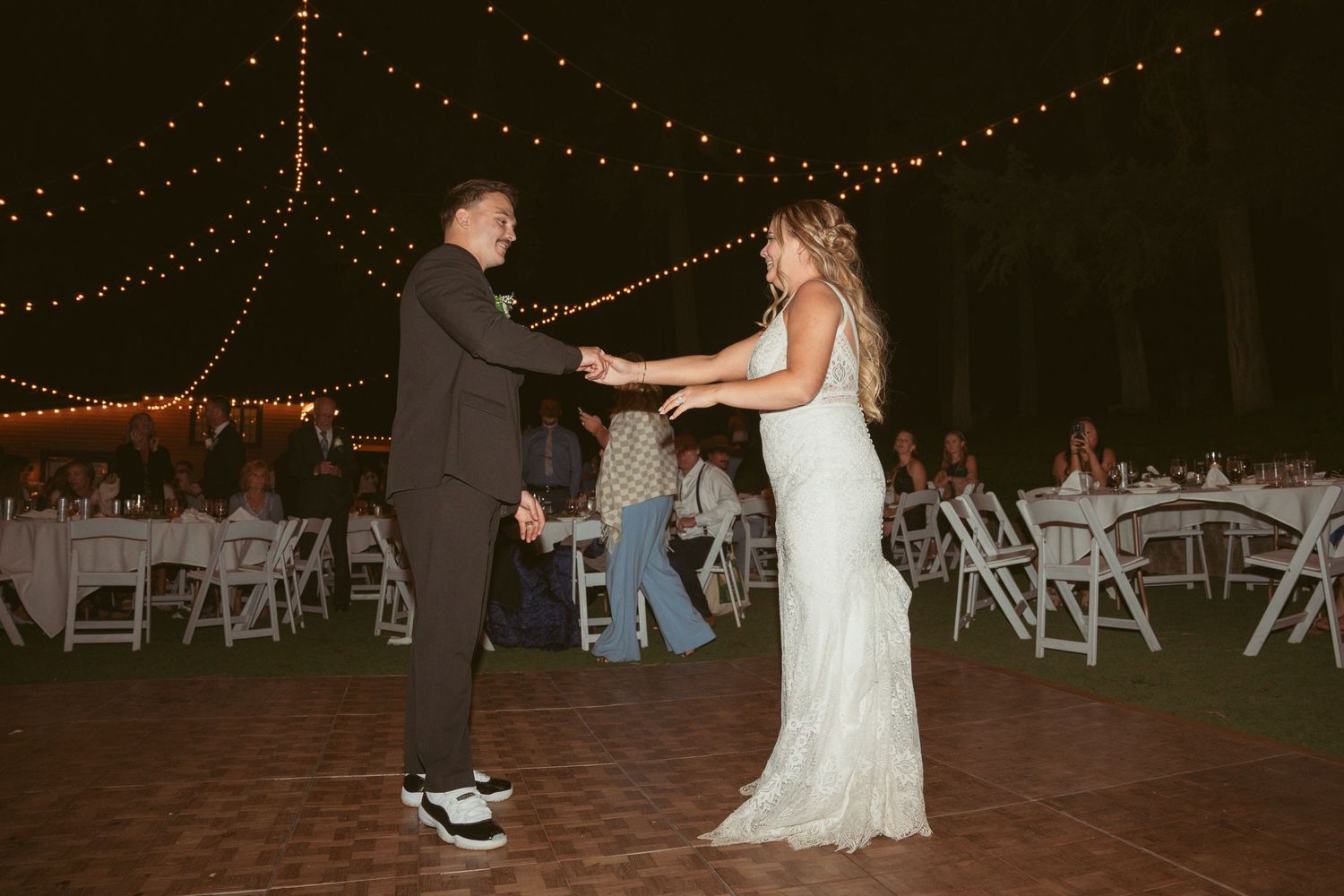 Couple shares their first dance on an outdoor dance floor under romantic string lighting.