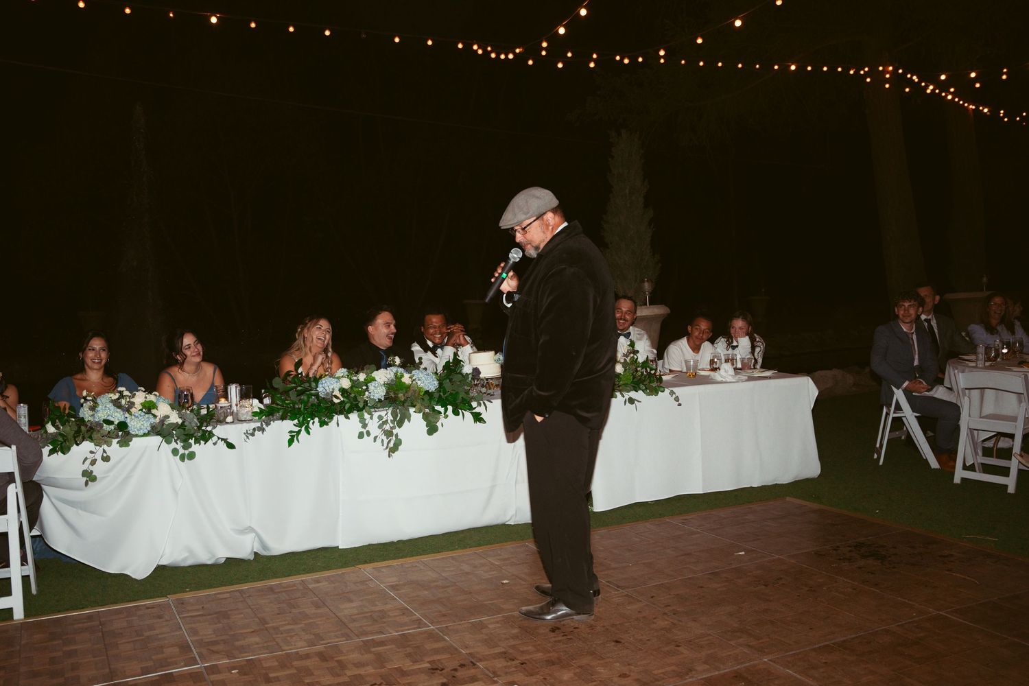 A panoramic view of a dimly lit outdoor wedding reception with string lights and white table settings at dusk.