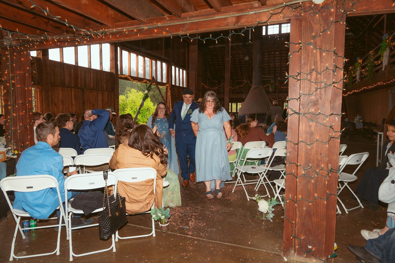 Wedding guests seated in folding chairs inside a rustic barn venue decorated with string lights.