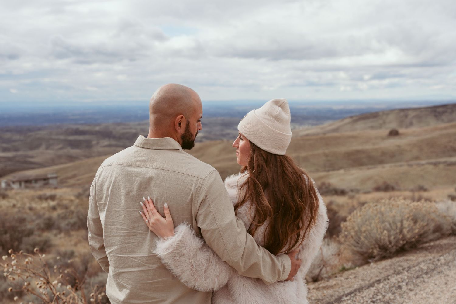 Couple sitting together on a scenic hillside overlooking a vast mountain landscape on an overcast day.