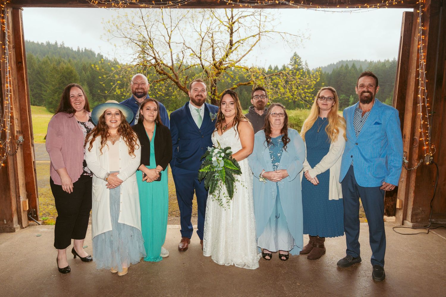 Wedding group in mix of blue and green attire poses under wooden arch with mountain view.