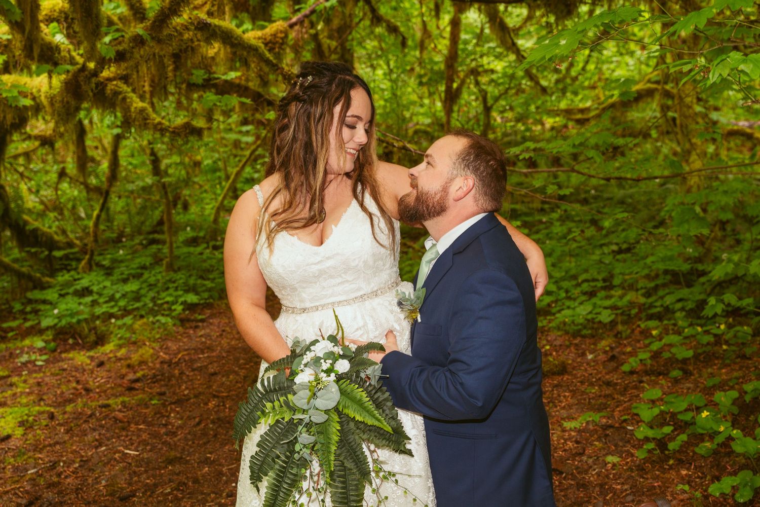 Wedding couple embraces under lush green forest canopy during photoshoot.