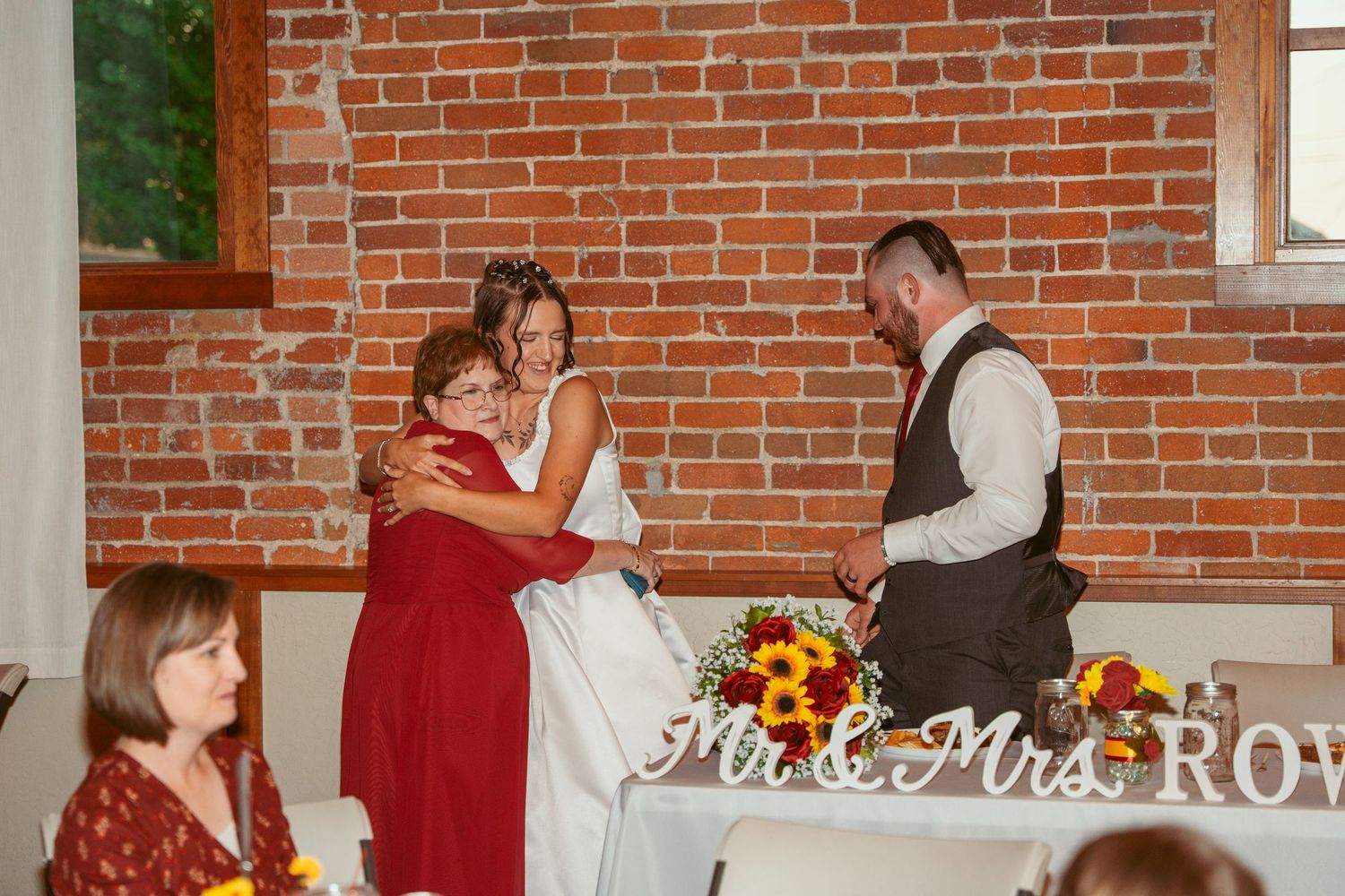 Wedding guests gather in a rustic brick-walled reception venue with sunflower decorations and white tablecloths.