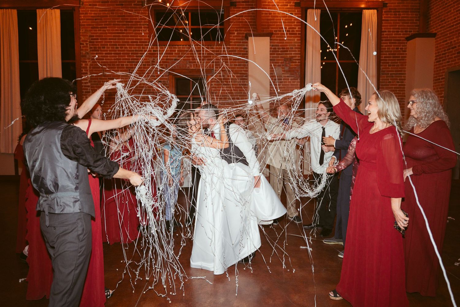 Wedding party throws streamers during celebration in venue with exposed brick walls and string lights.