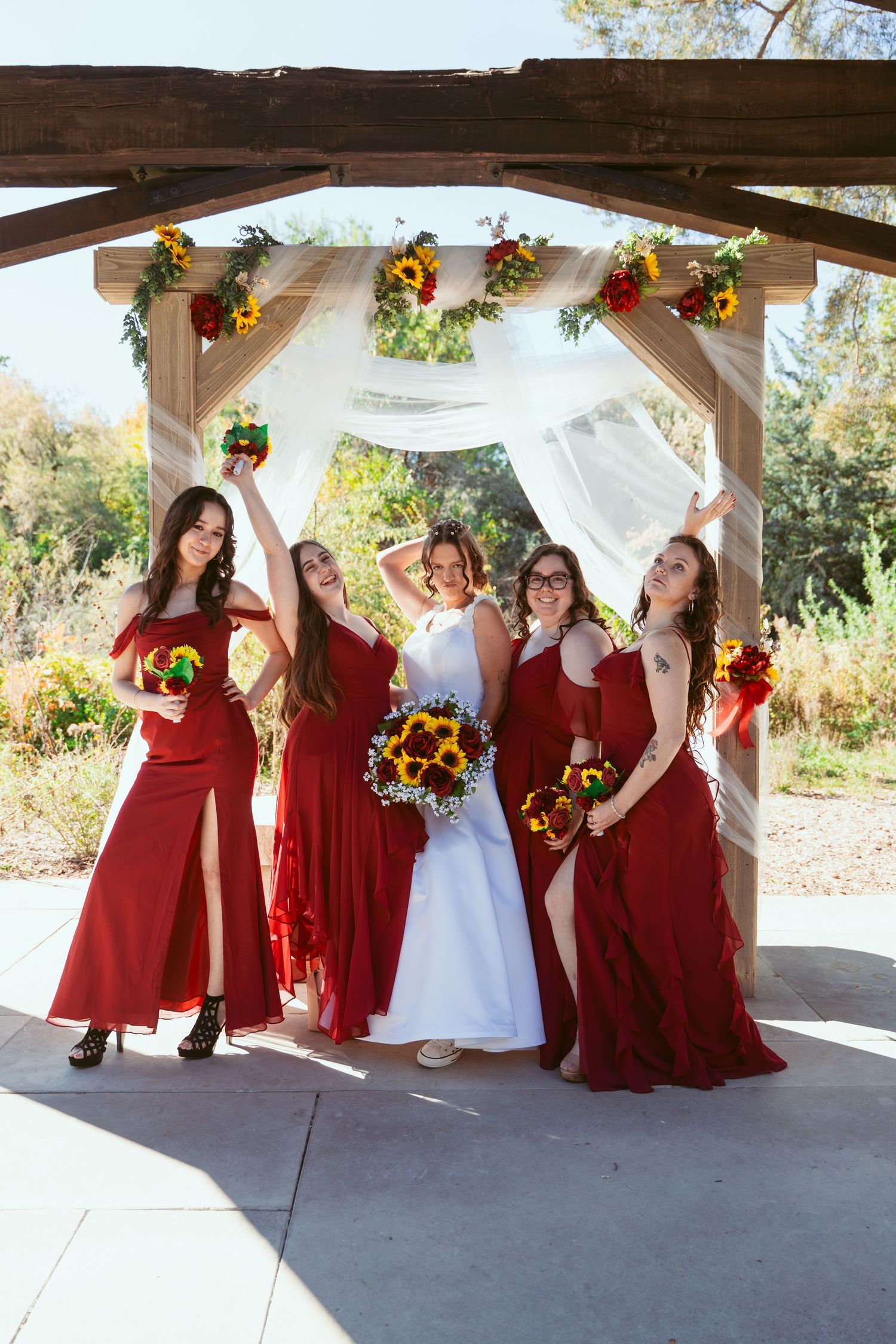 Bridal party in crimson dresses poses playfully with sunflowers under white draped pavilion.