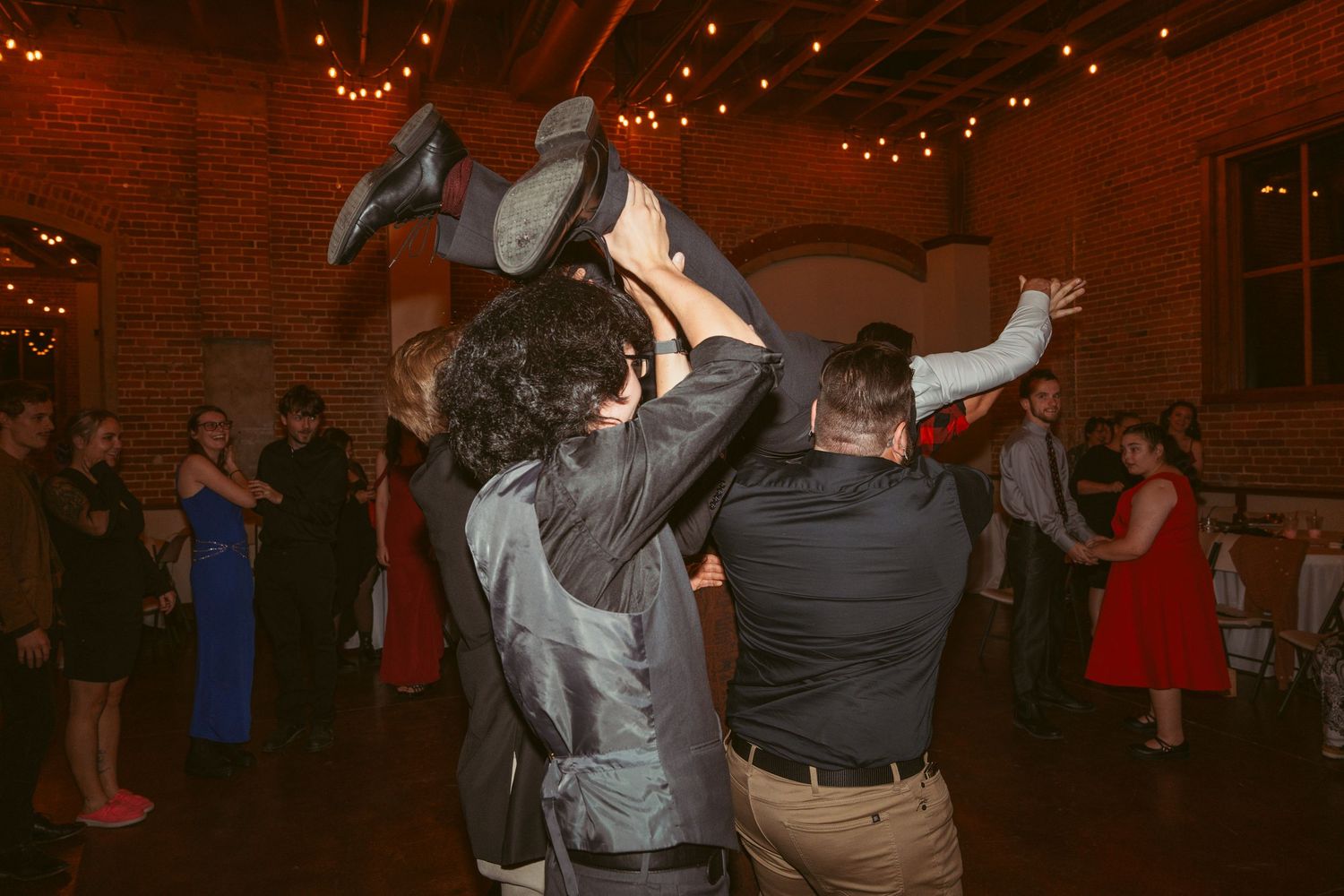 Dancers performing acrobatic lifts during a celebration in an industrial venue with string lights.