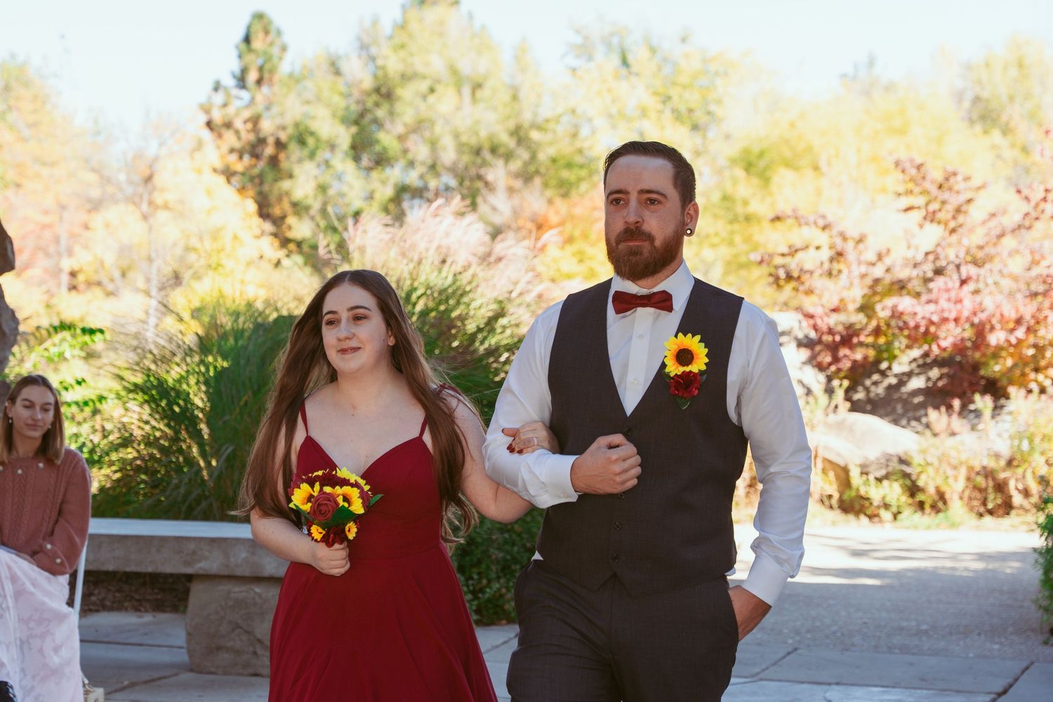 Wedding party members in red and black attire walk arm-in-arm down an outdoor stone path during autumn.