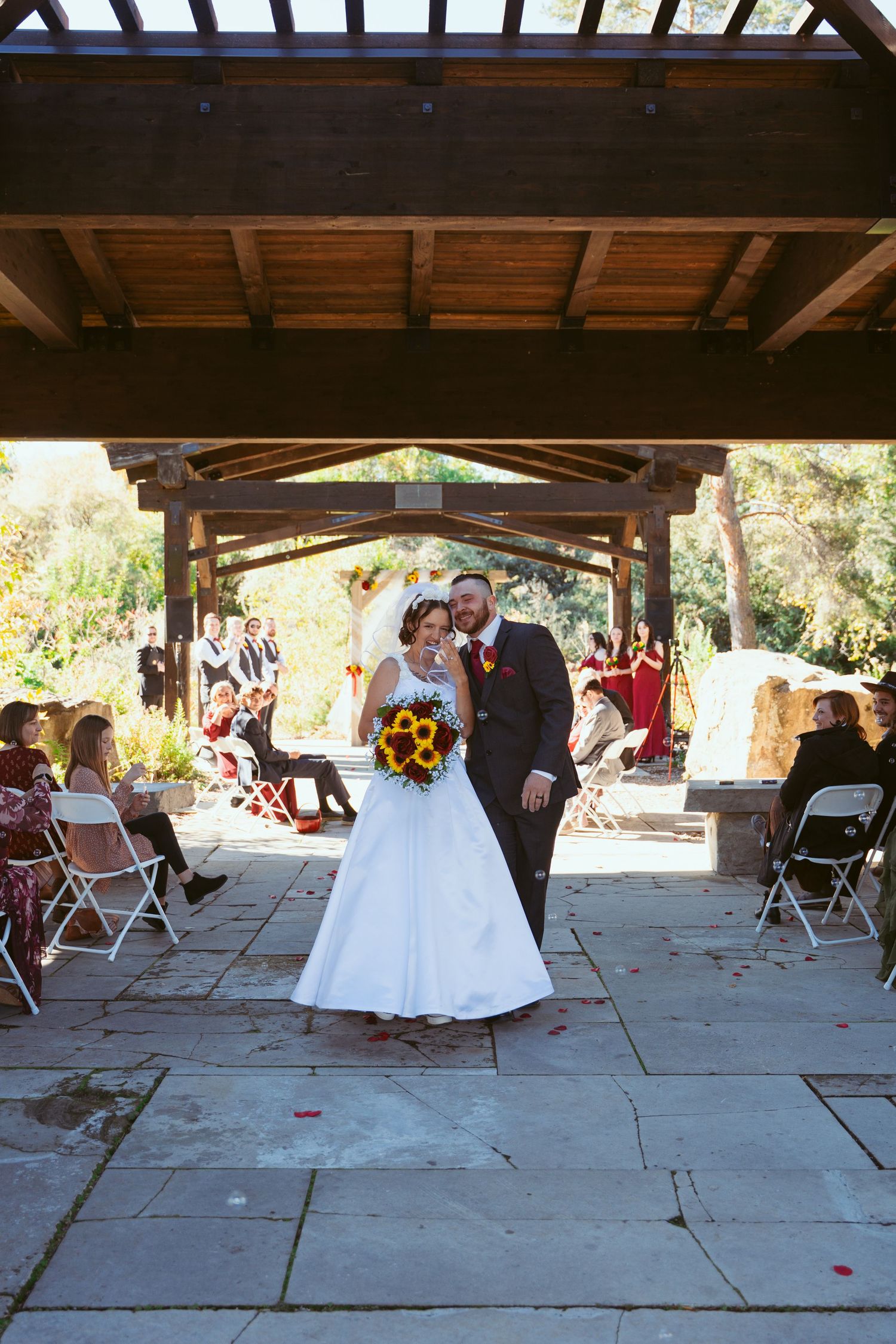Wedding celebration in an outdoor covered venue with wooden beams and stone columns.