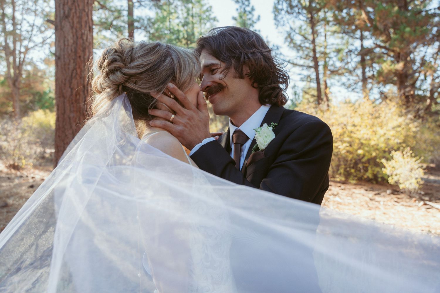 Wedding couple embracing outdoors with pine trees and flowing veil in background.