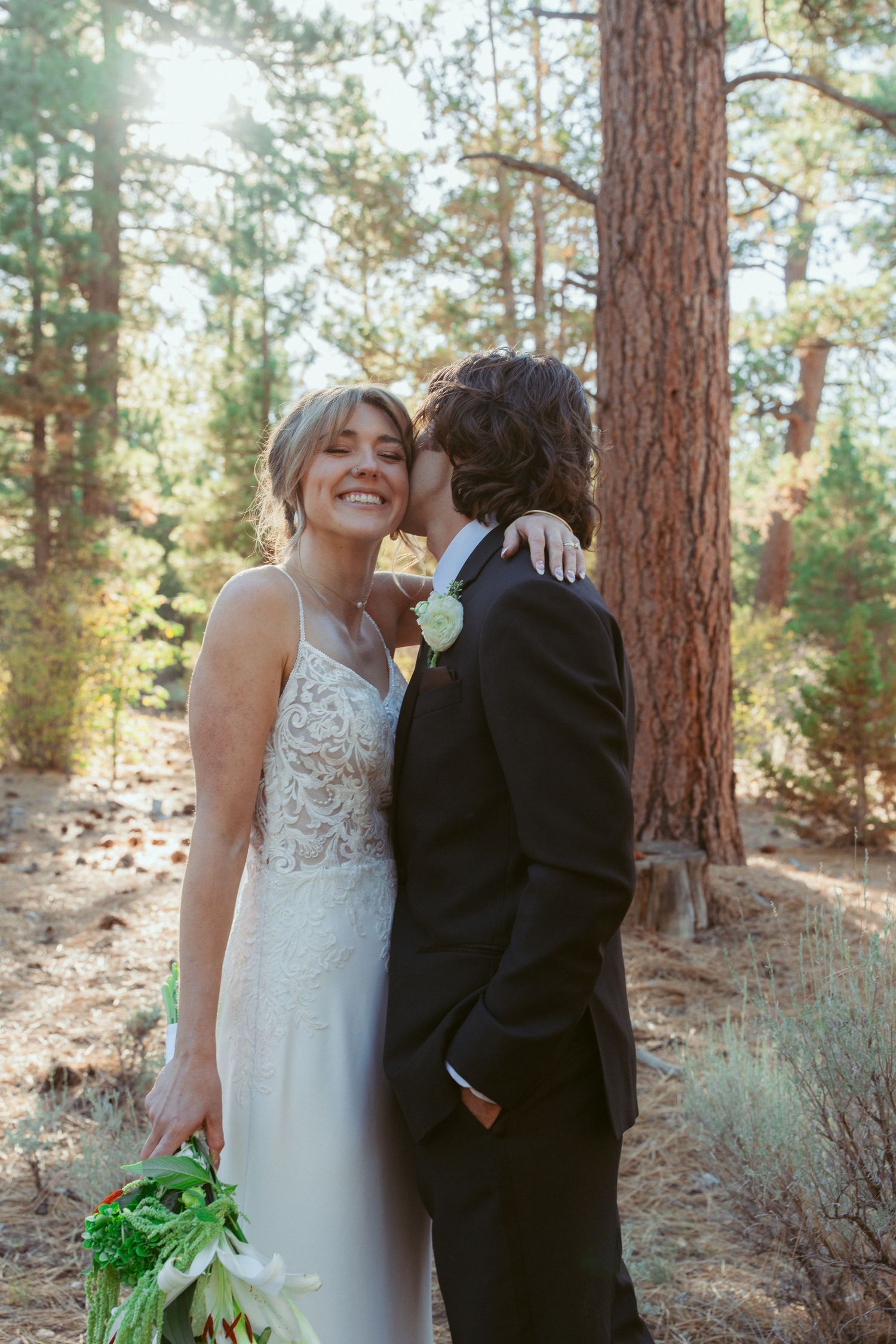 A romantic wedding couple shares an intimate moment among tall pine trees in a forest setting.