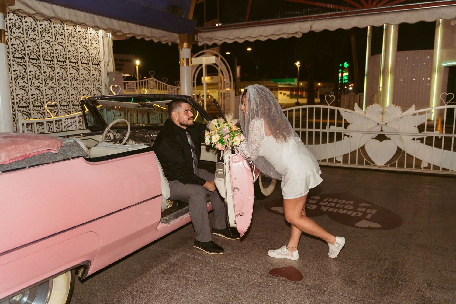Bride and groom pose next to a vintage pink car during their nighttime wedding celebration.