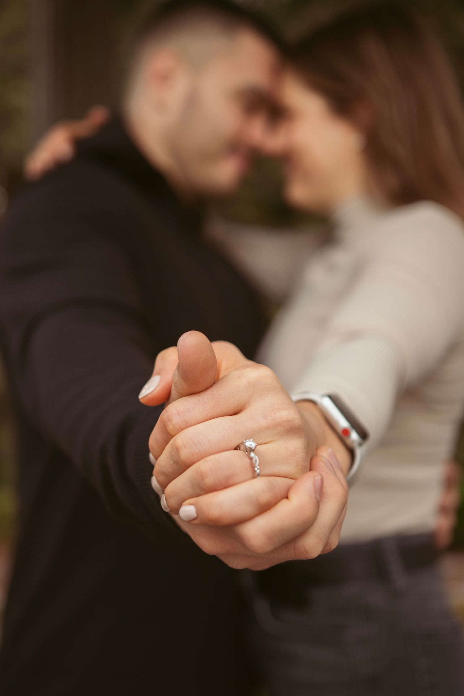 Close-up of clasped hands showing an engagement ring with a blurred couple in the background.