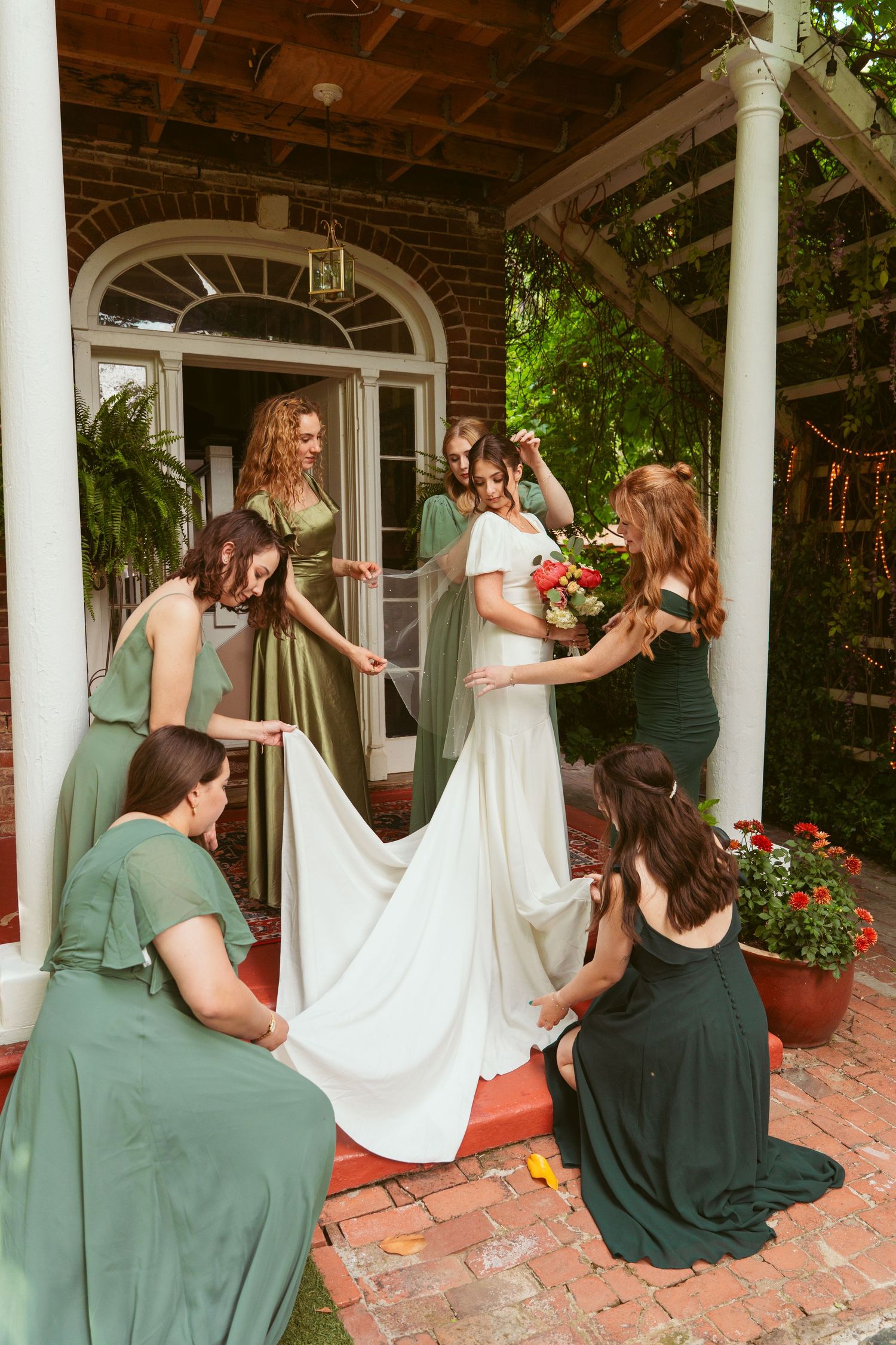 Bridesmaids helping arrange a wedding dress train on a covered brick porch.