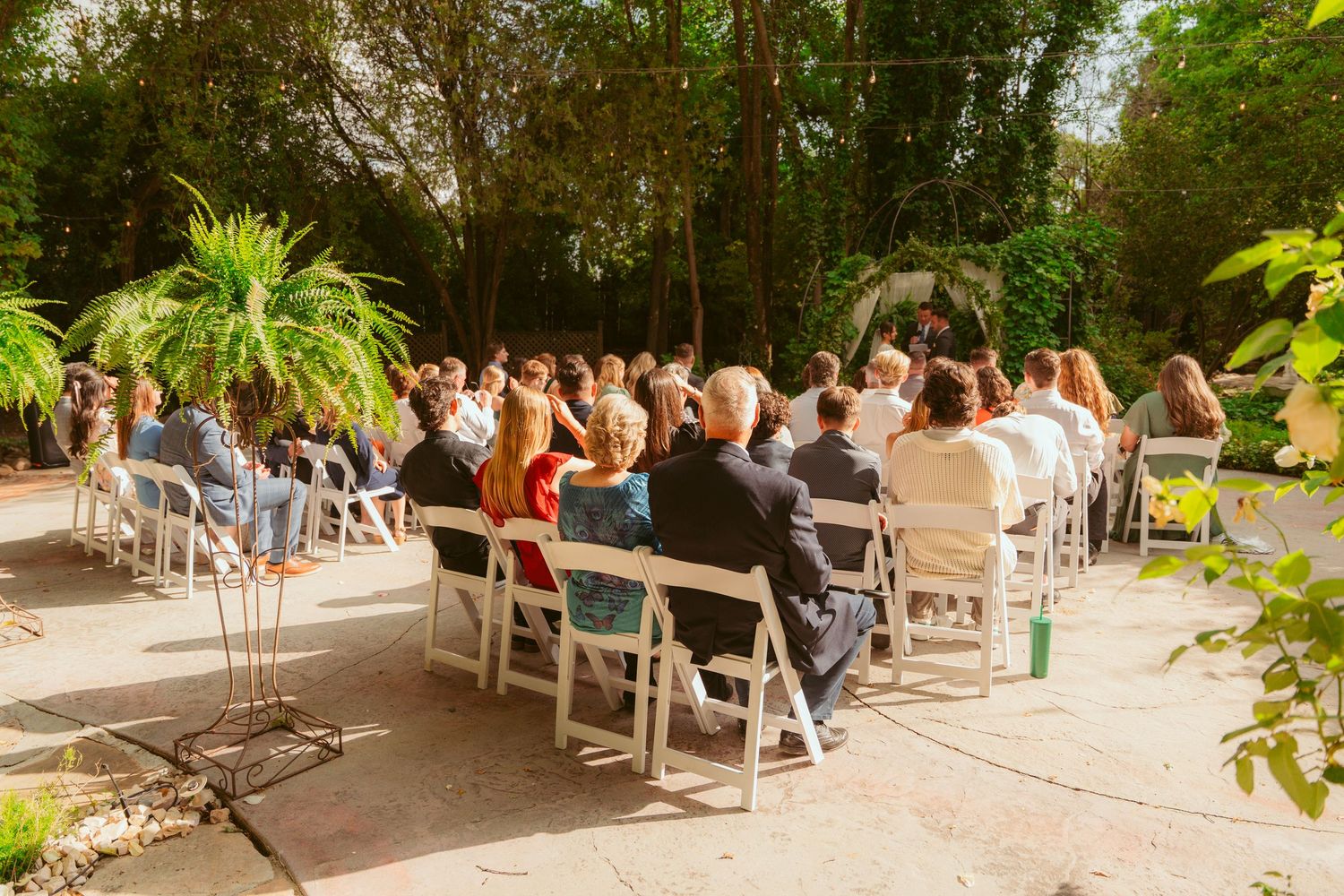 Panoramic view of wedding guests seated in white chairs at an outdoor garden ceremony.