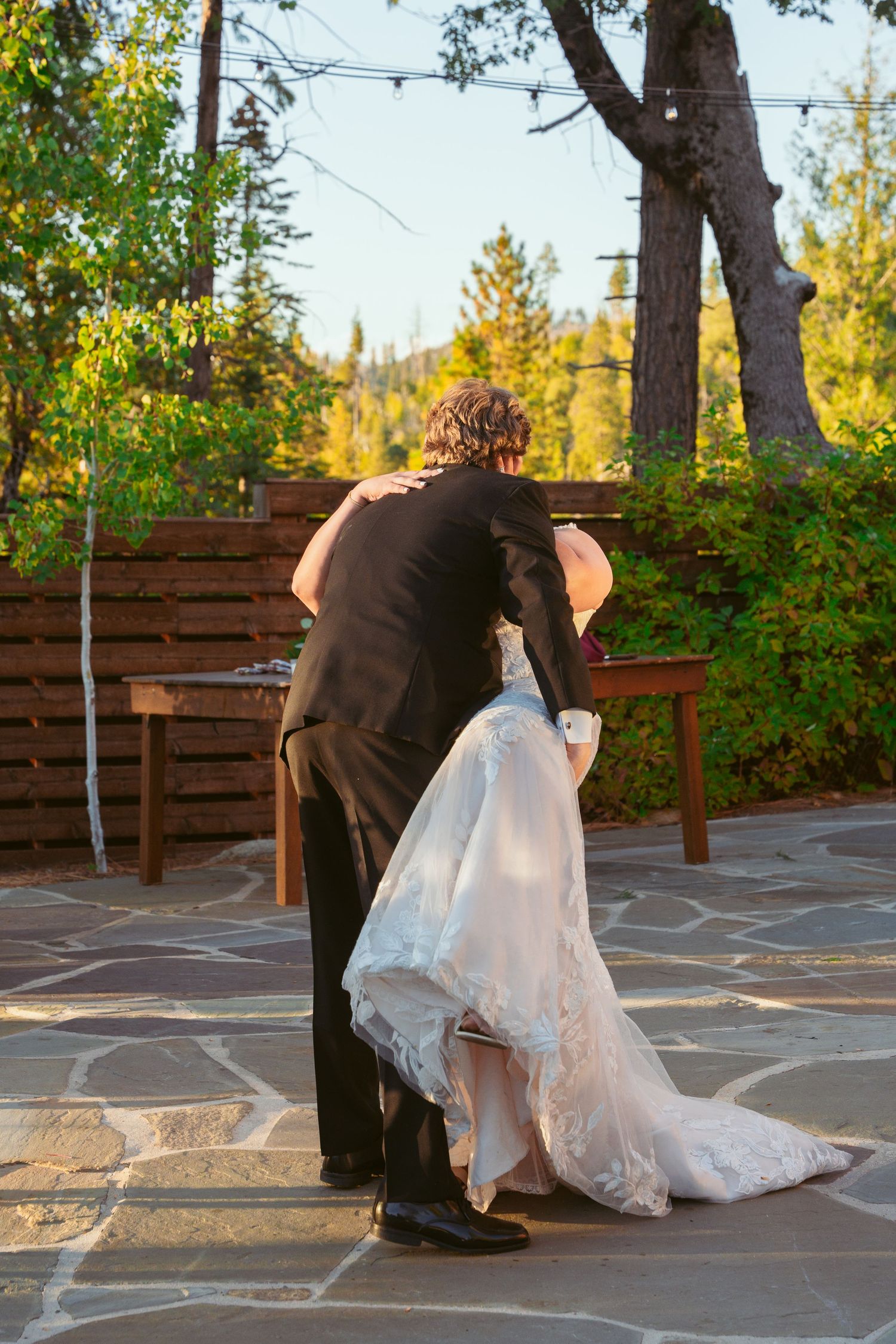 Romantic dance dip sequence on stone patio with wooden benches and forest backdrop.