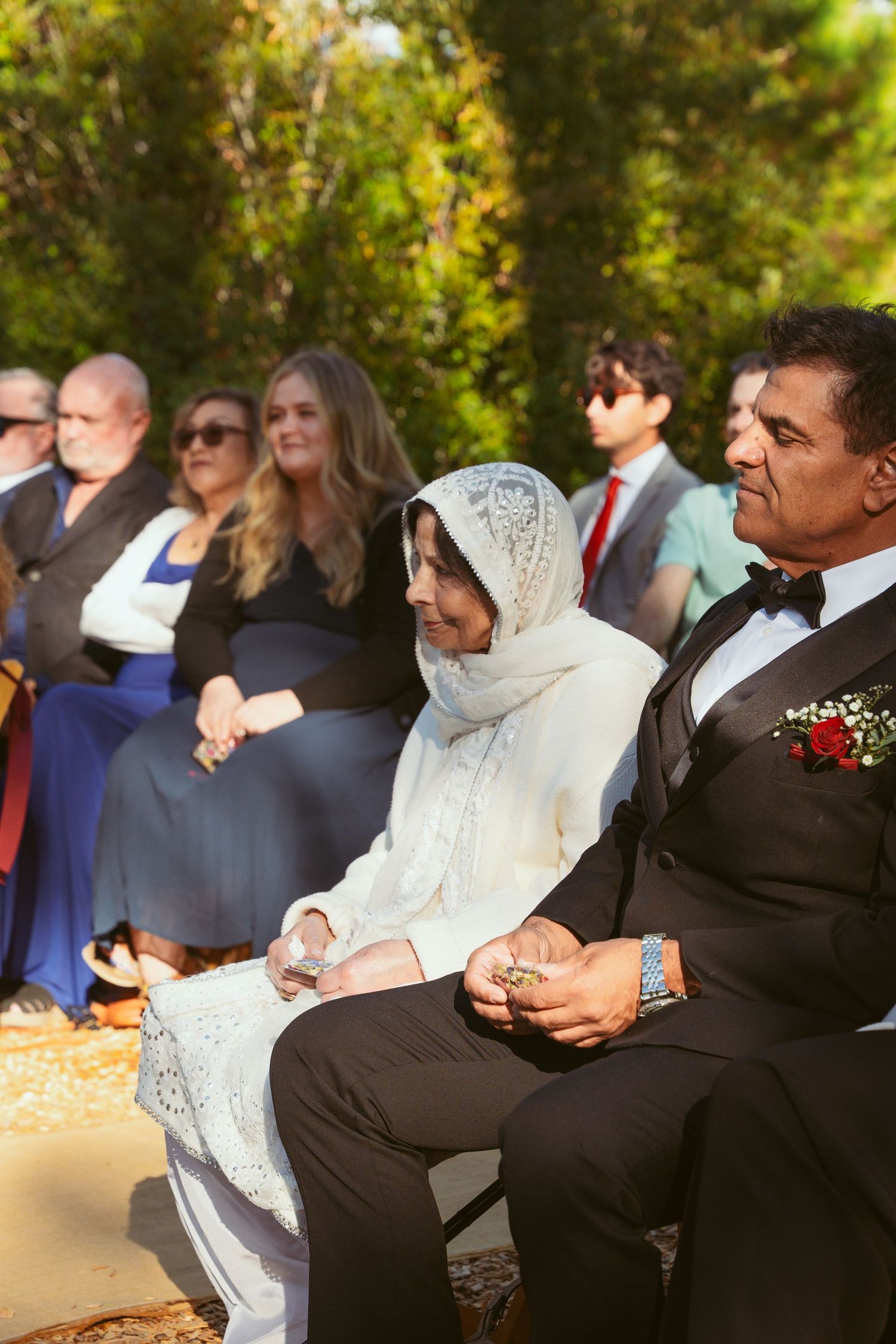 Wedding guests seated outdoors watching ceremony in a wooded setting with filtered sunlight.