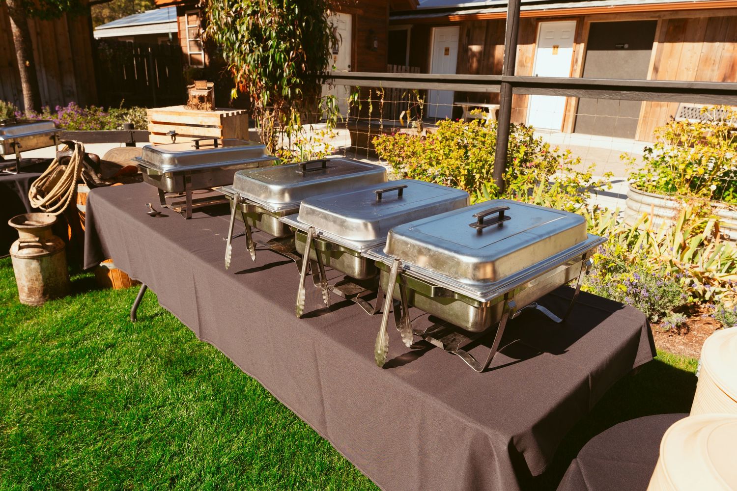 Row of stainless steel chafing dishes set up on brown tablecloth for outdoor catering service.