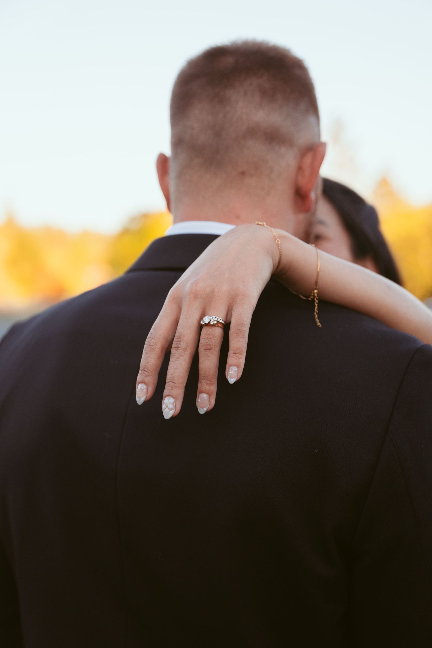 Close-up of hands with wedding rings embracing against a black suit jacket at sunset.