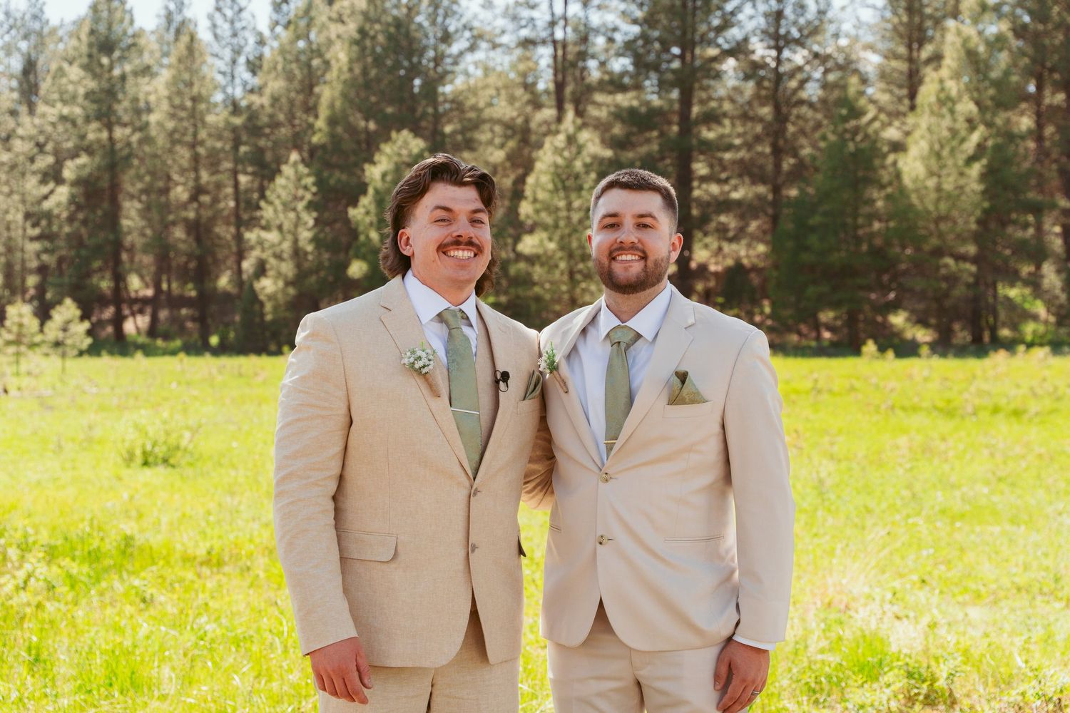 Wedding party members in light colored suits smile and pose together in an outdoor meadow setting.