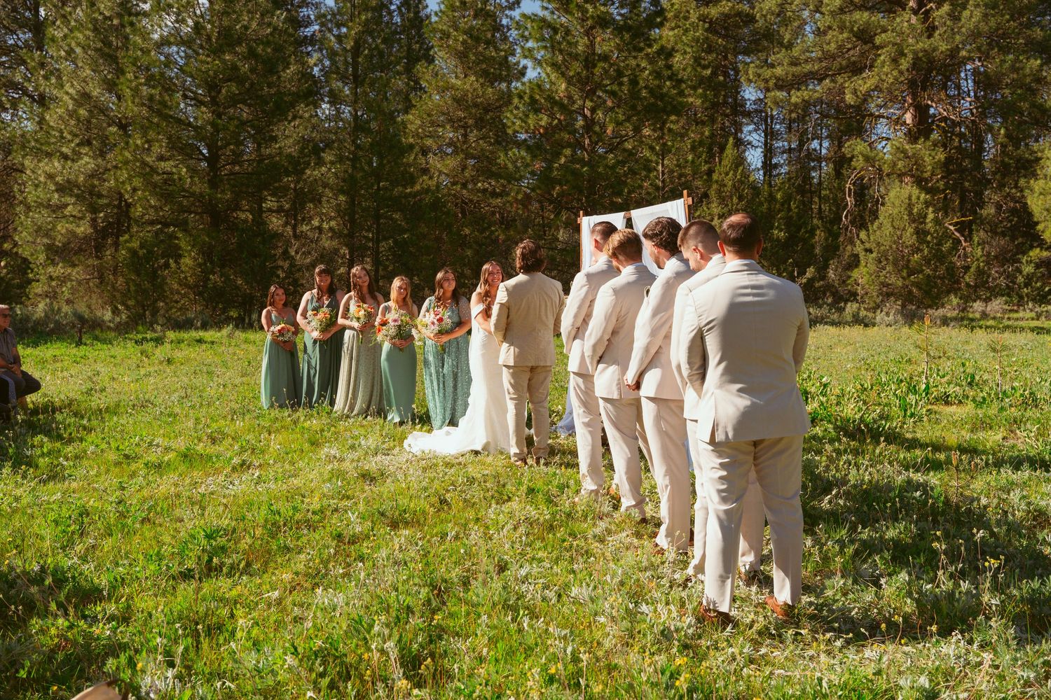 Wedding party lines up in sage green dresses and beige suits against forest backdrop.