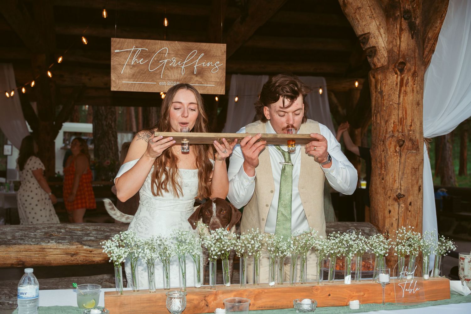 Wedding party members take shots at a rustic barn reception with wooden beams and string lights.
