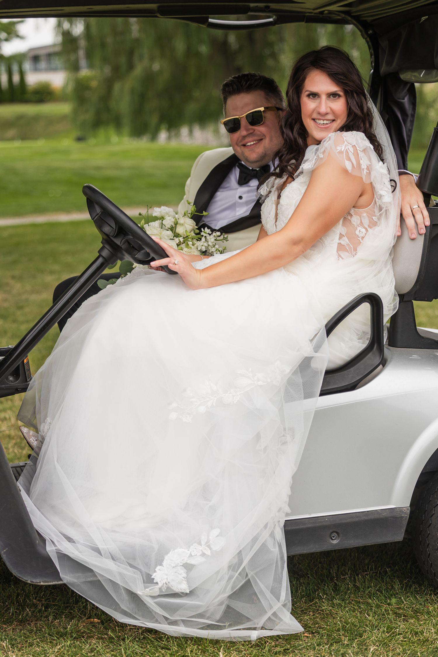 Smiling bride and groom riding together in a white golf cart on a green lawn.