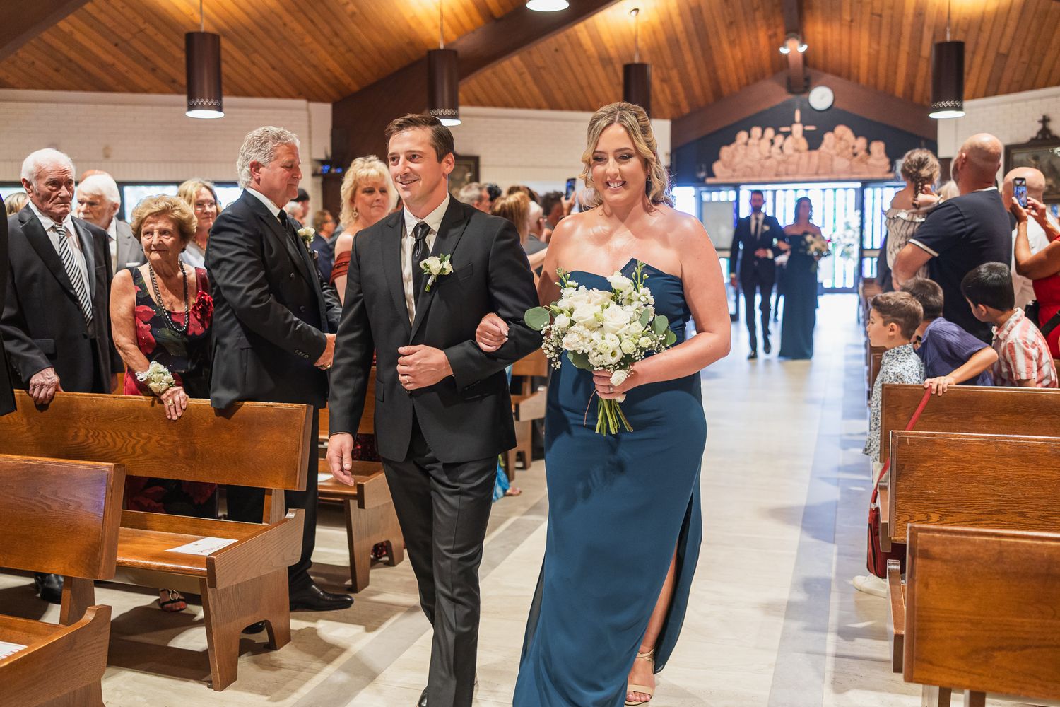 Groomsman escorts bridesmaid in navy dress down church aisle holding white bouquet at wedding.