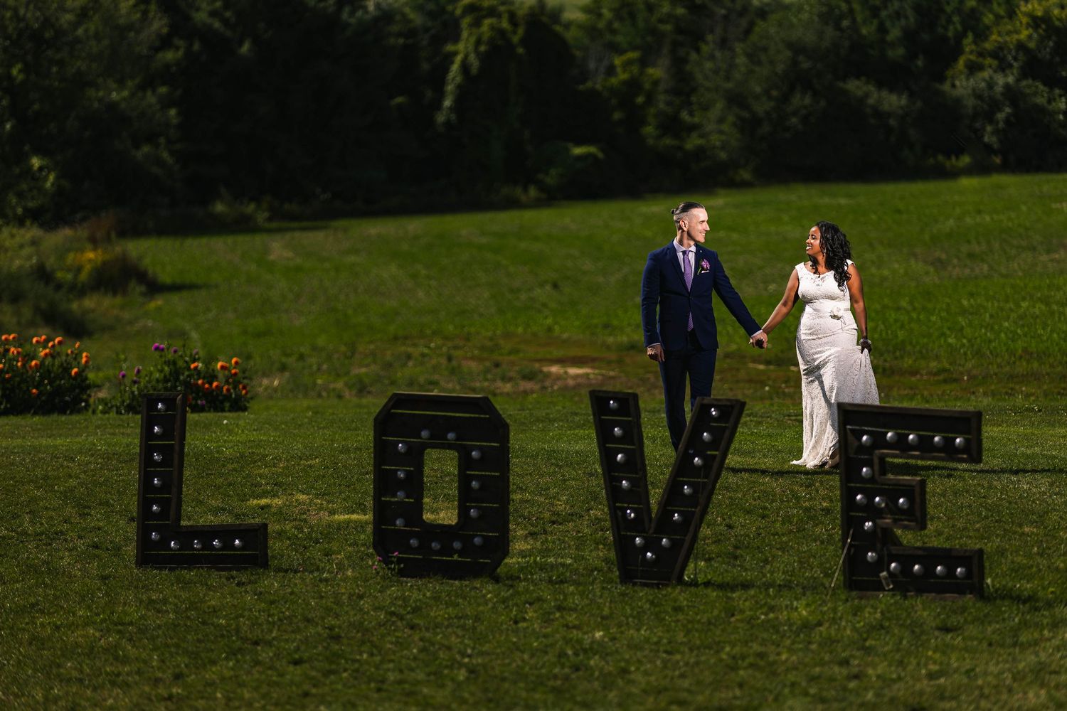Wedding couple poses near large illuminated LOVE letters on grassy lawn.