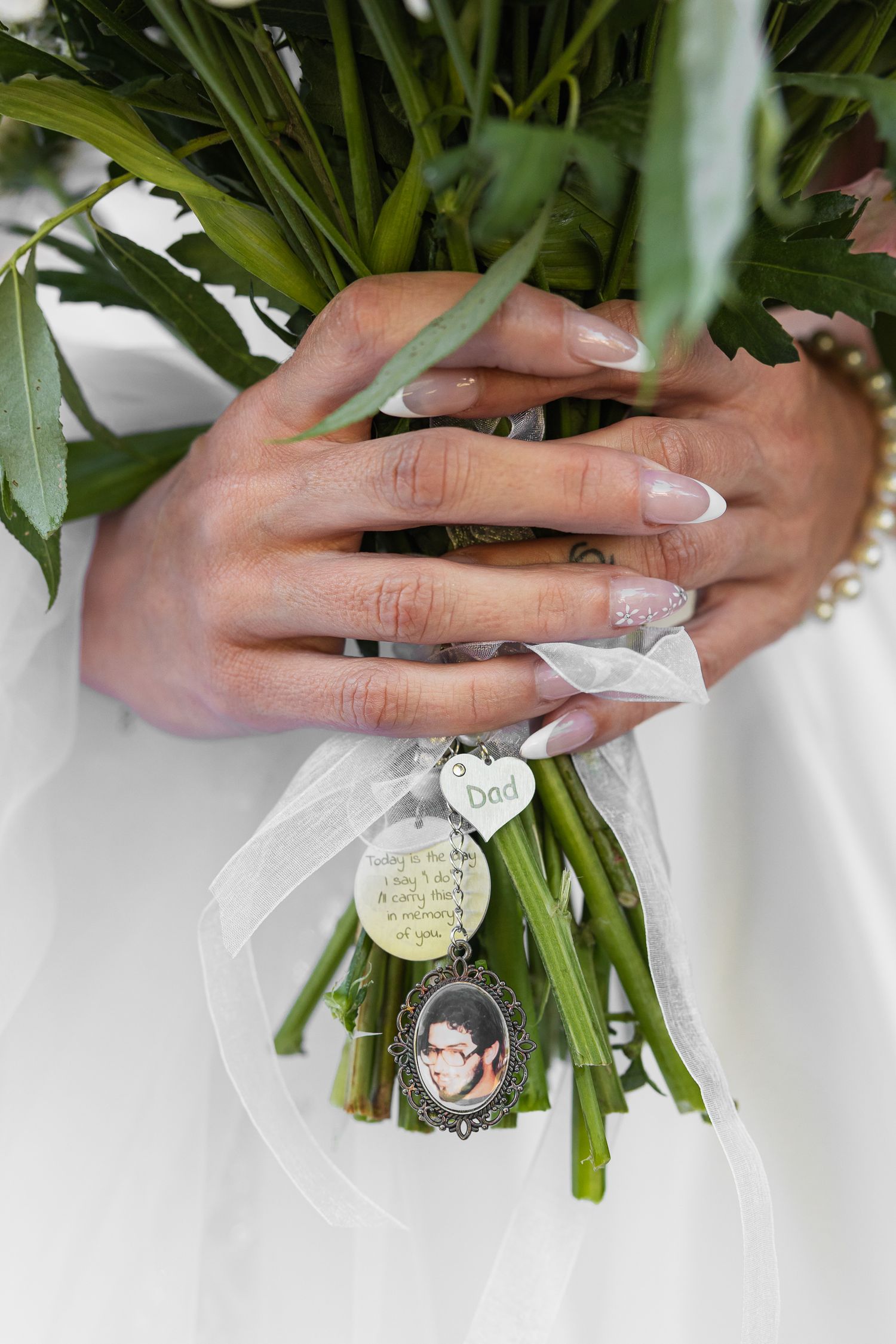 Bride holding white wedding bouquet with memorial charms including a photo pendant and heart charm with French manicured nails.