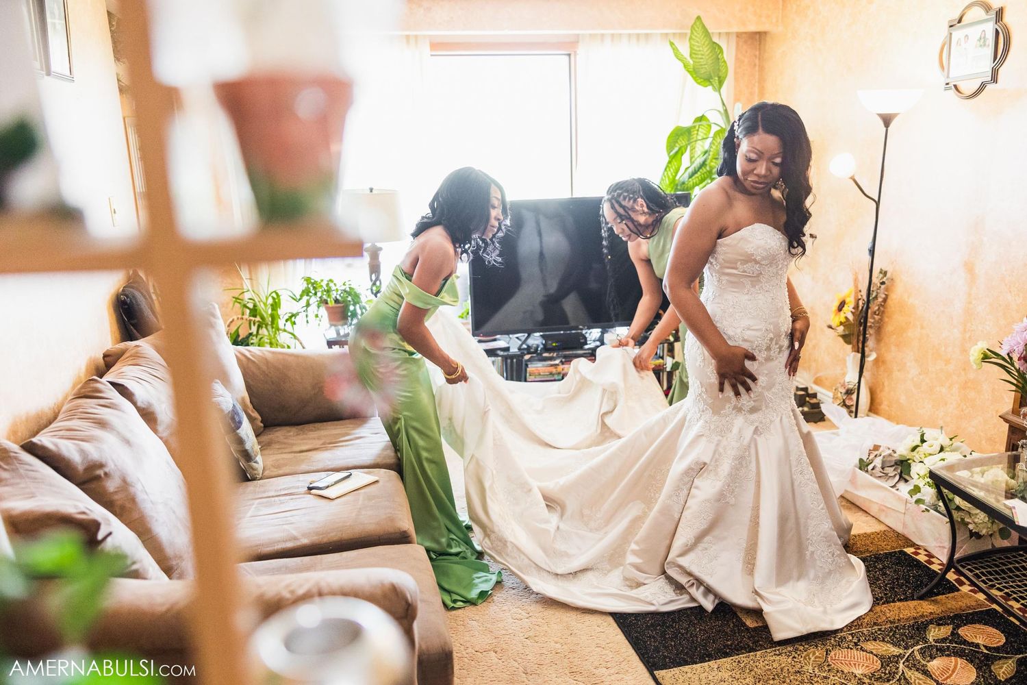 Wedding party assists with arranging the train of an elegant white mermaid gown in a sunlit room.
