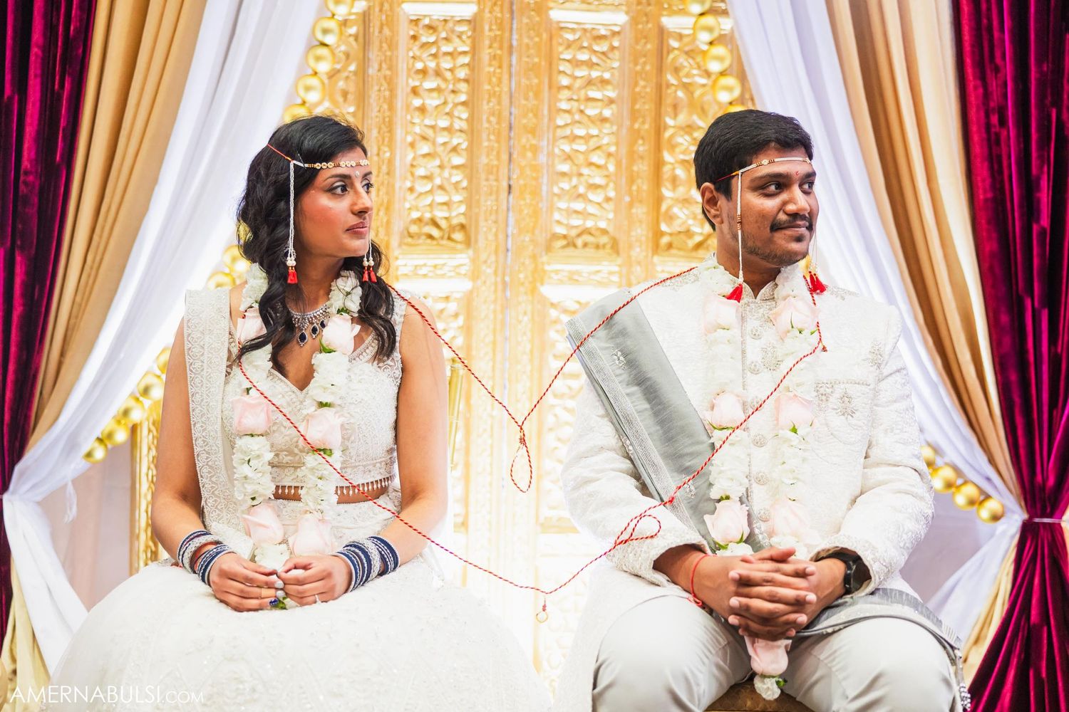 Wedding couple seated on white chairs during traditional ceremony with ornate gold backdrop.