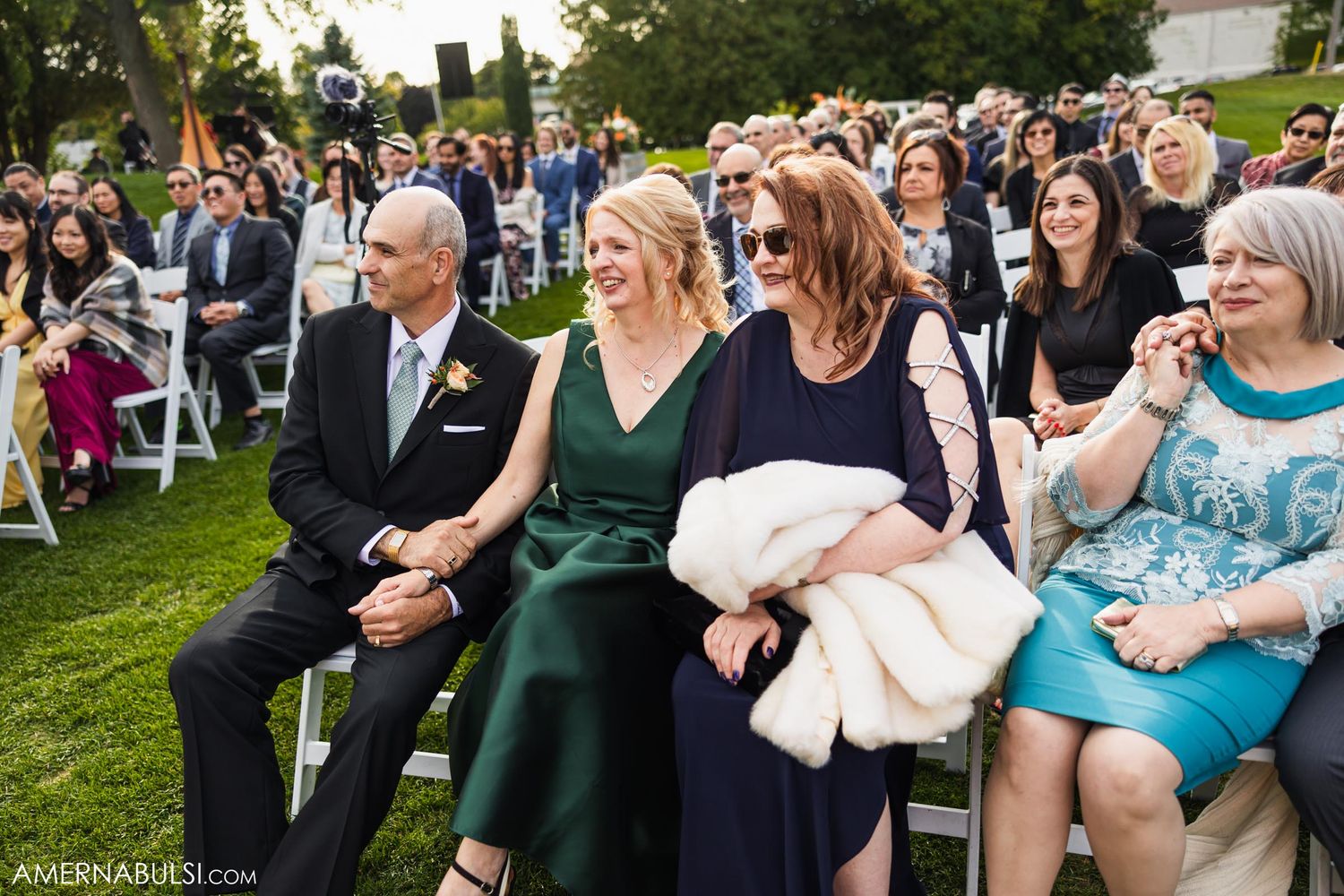 Wedding guests seated on chairs watching an outdoor ceremony with greenery in the background.