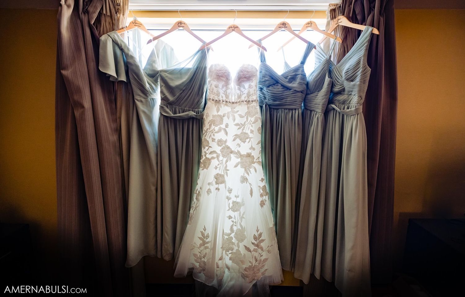 Wedding dress and bridesmaid dresses hanging in window with soft natural light.