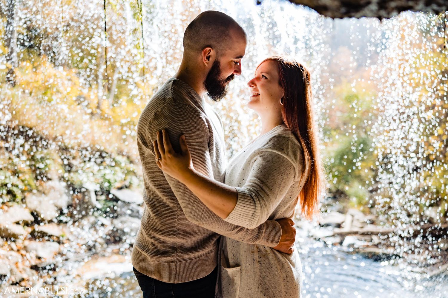 Romantic couple embracing under waterfall with sunlit spray.