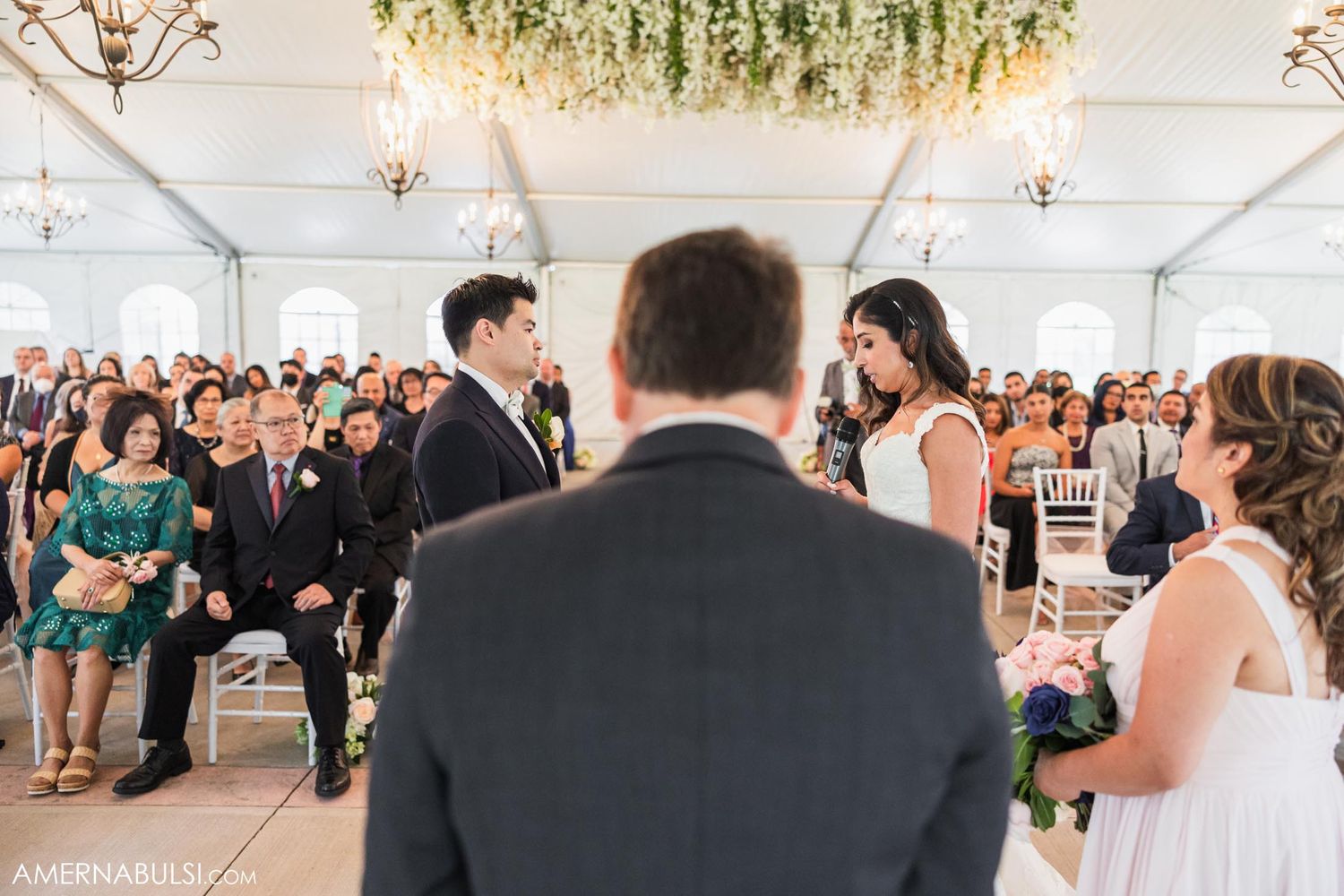 Wedding ceremony in a light-filled tent with hanging greenery and chandeliers.