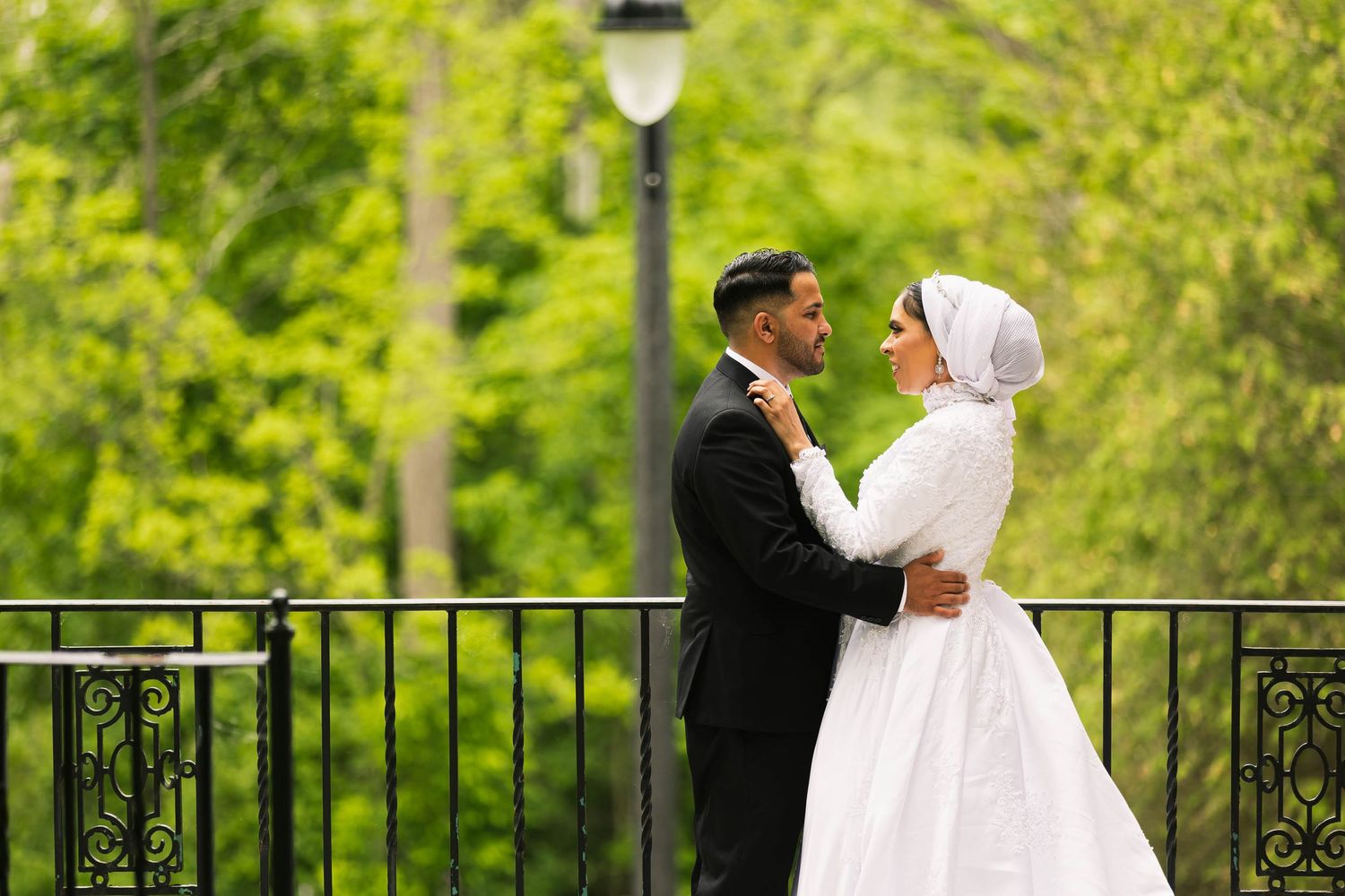 Couple in formal wedding attire embracing outdoors near decorative railing and lamp post.