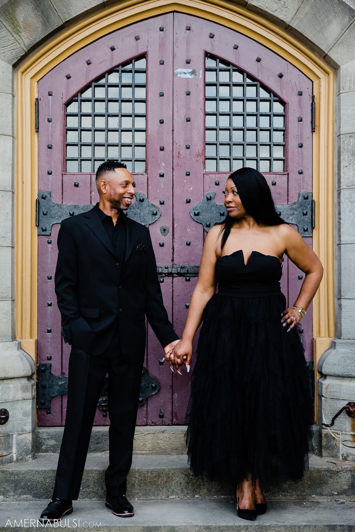 Couple in matching black outfits stands before decorative pink cathedral doors.