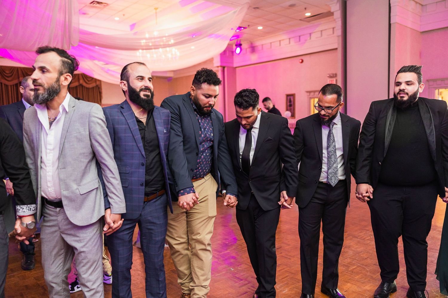 Group of men in formal attire walking together at a wedding reception with pink ambient lighting.