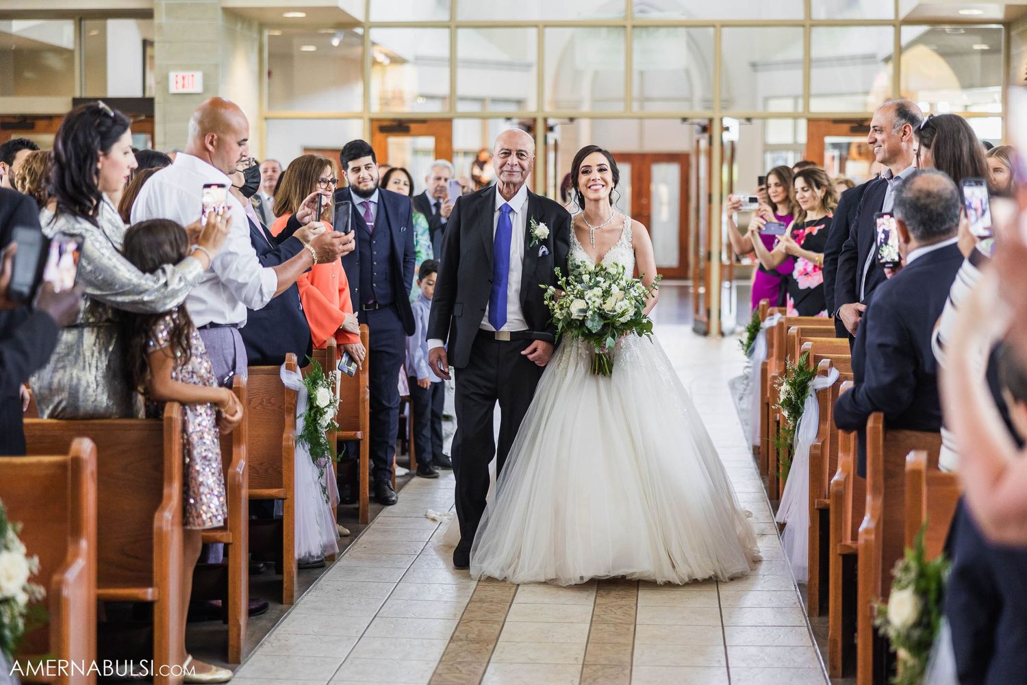 Wedding guests stand in church pews as a couple processes down the aisle decorated with floral arrangements.