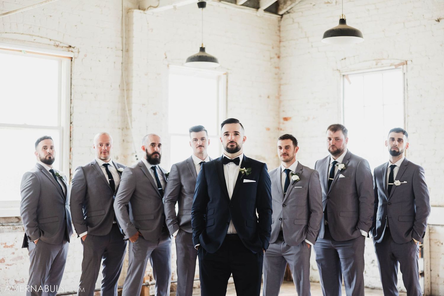 Groomsmen in coordinated grey suits stand with groom in black tux in bright white industrial space.
