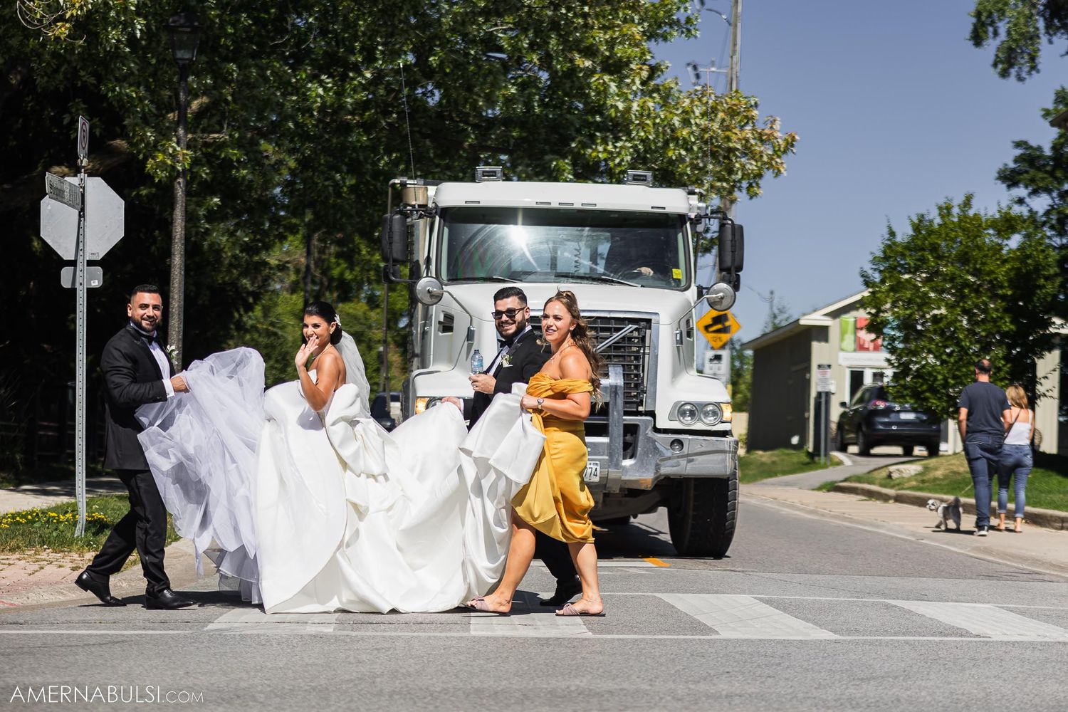 Wedding party helps carry bride's dress train near white truck on sunny street.
