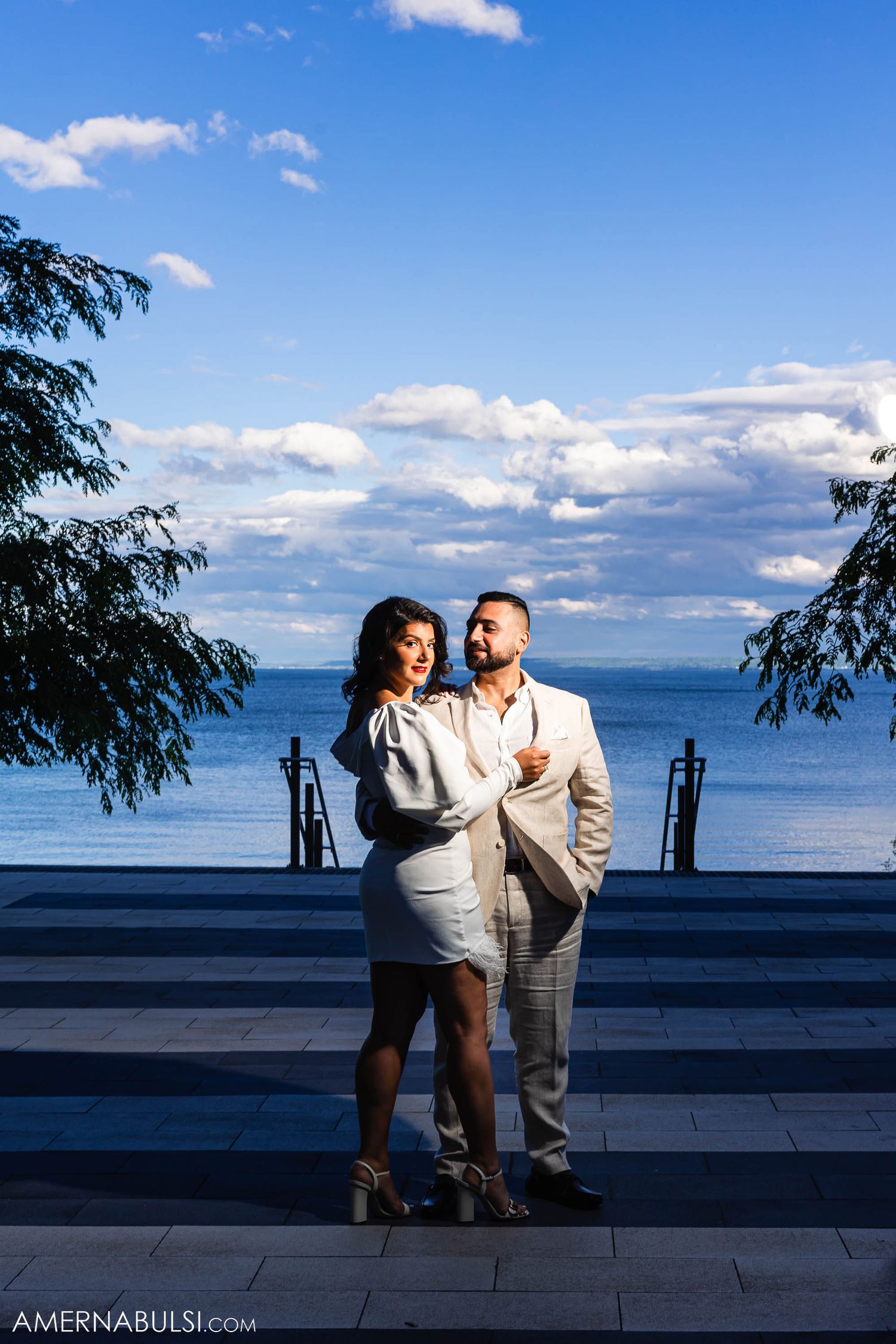 A couple in formal attire poses against a scenic waterfront backdrop with blue skies.
